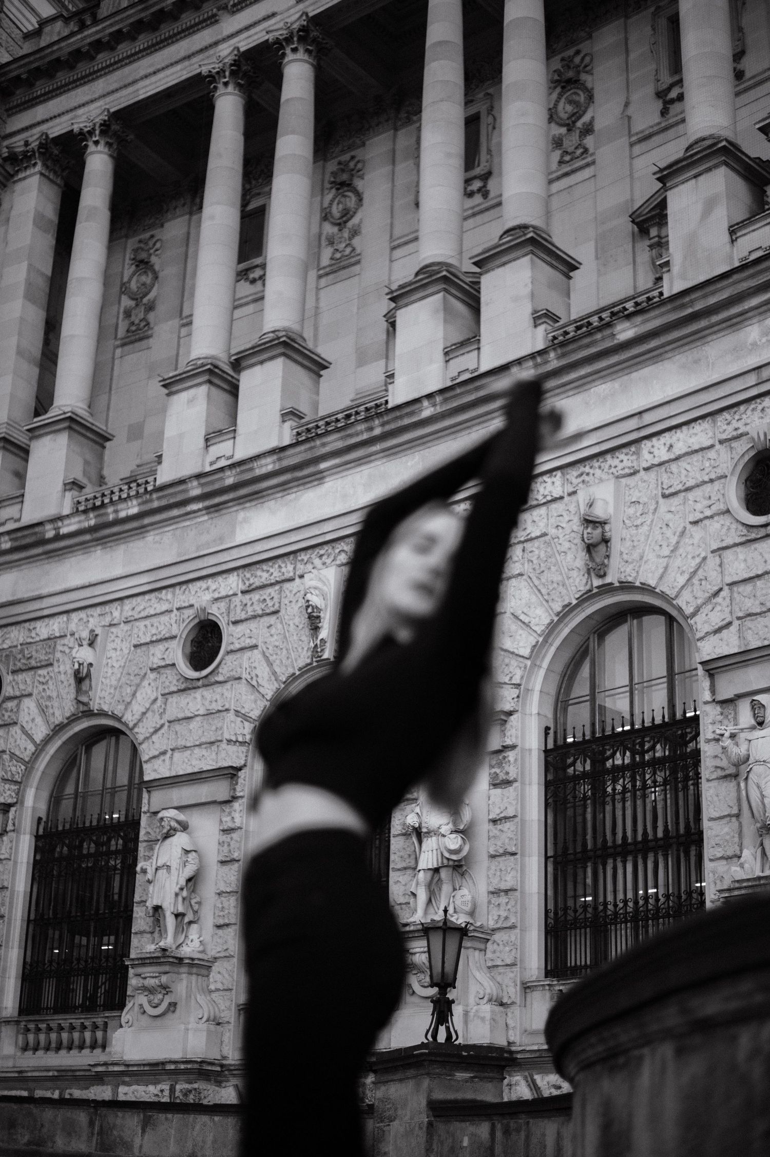 Black and white photo of woman in black outfit stretching arms up before grand neoclassical building.
