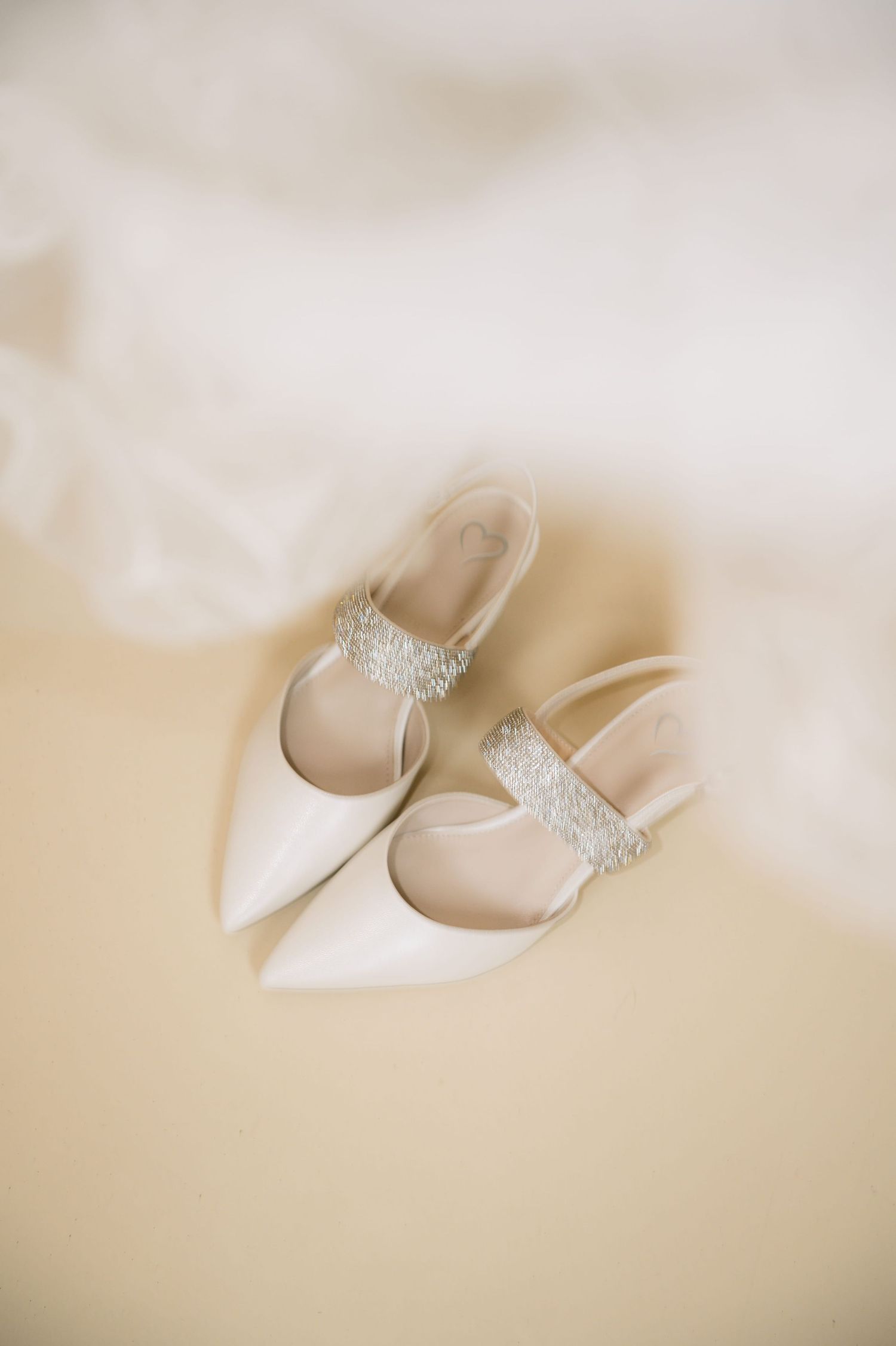 Elegant white pointed-toe bridal heels with crystal embellished ankle straps on soft fabric background.