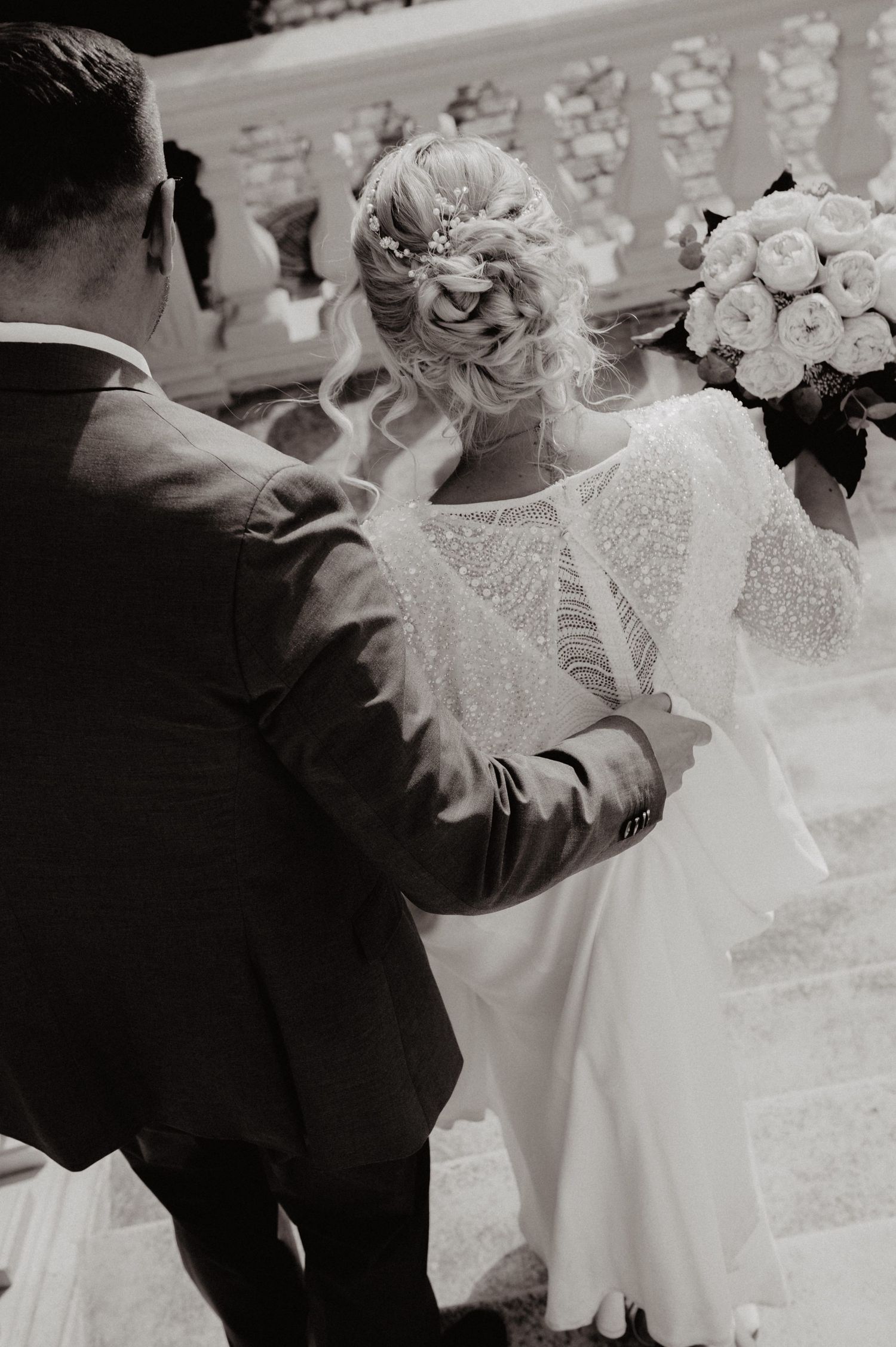 Black and white photo of bride with elegant updo holding bouquet while groom adjusts her beaded dress.
