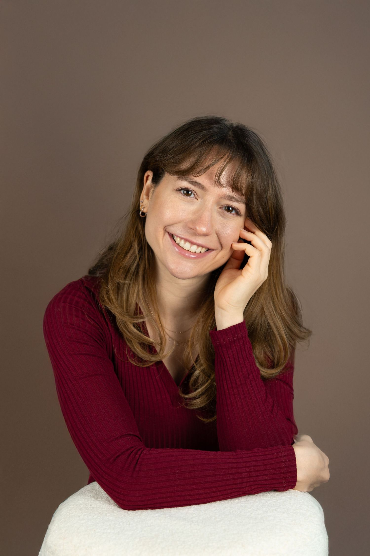 Smiling young woman with brown hair wearing a red ribbed sweater, posing with hand on cheek against brown background.