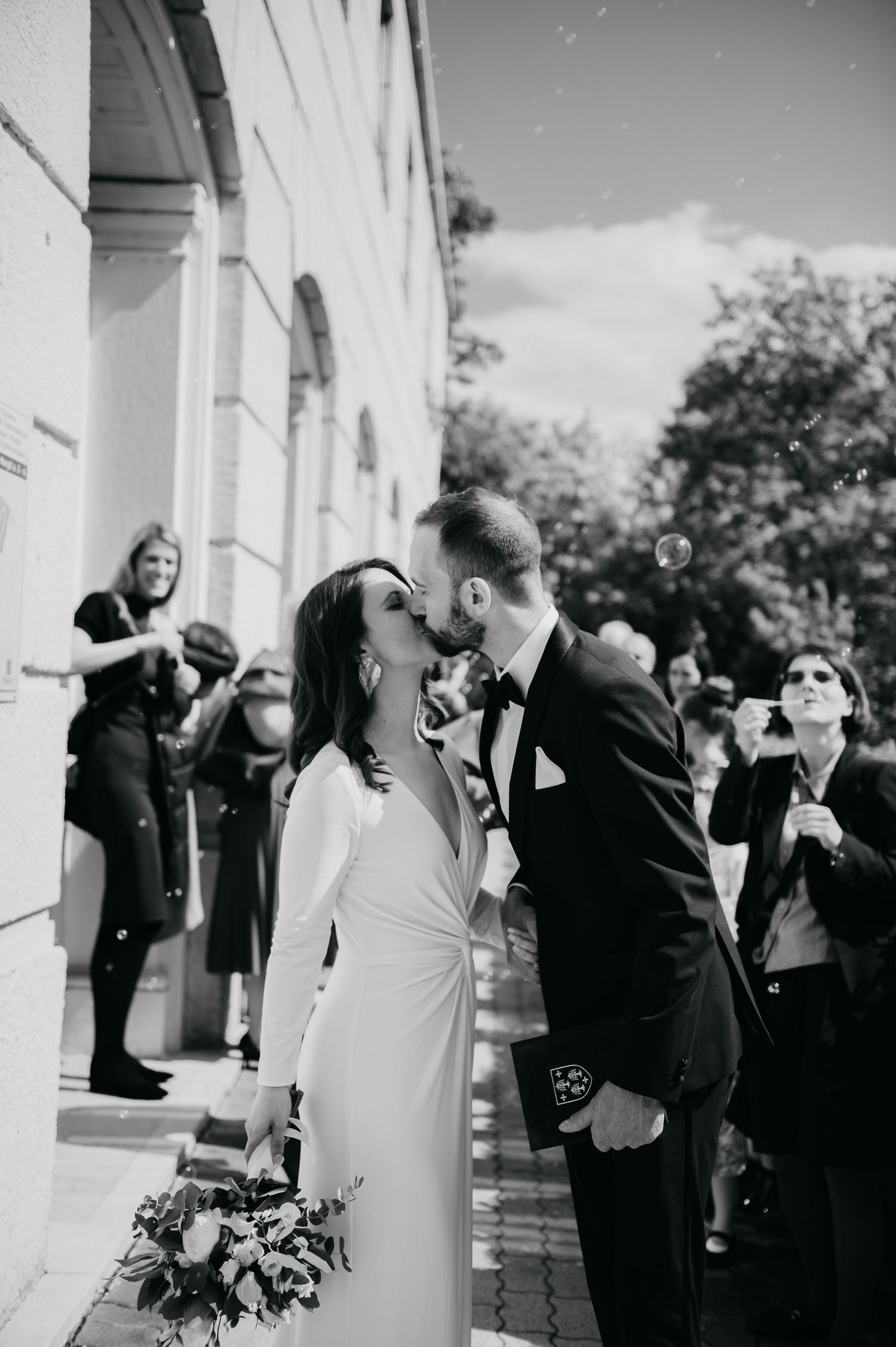 Black and white photo of a bride and groom kissing outside a church, surrounded by guests blowing bubbles.