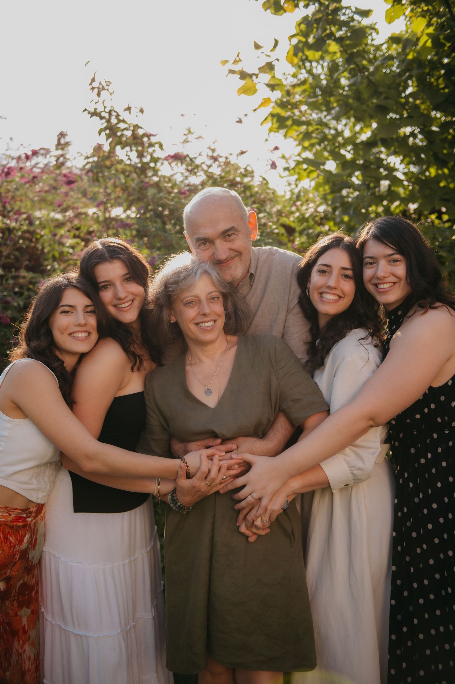 A joyful family of six poses together outdoors, smiling and holding hands in a sunlit garden with lush green trees.