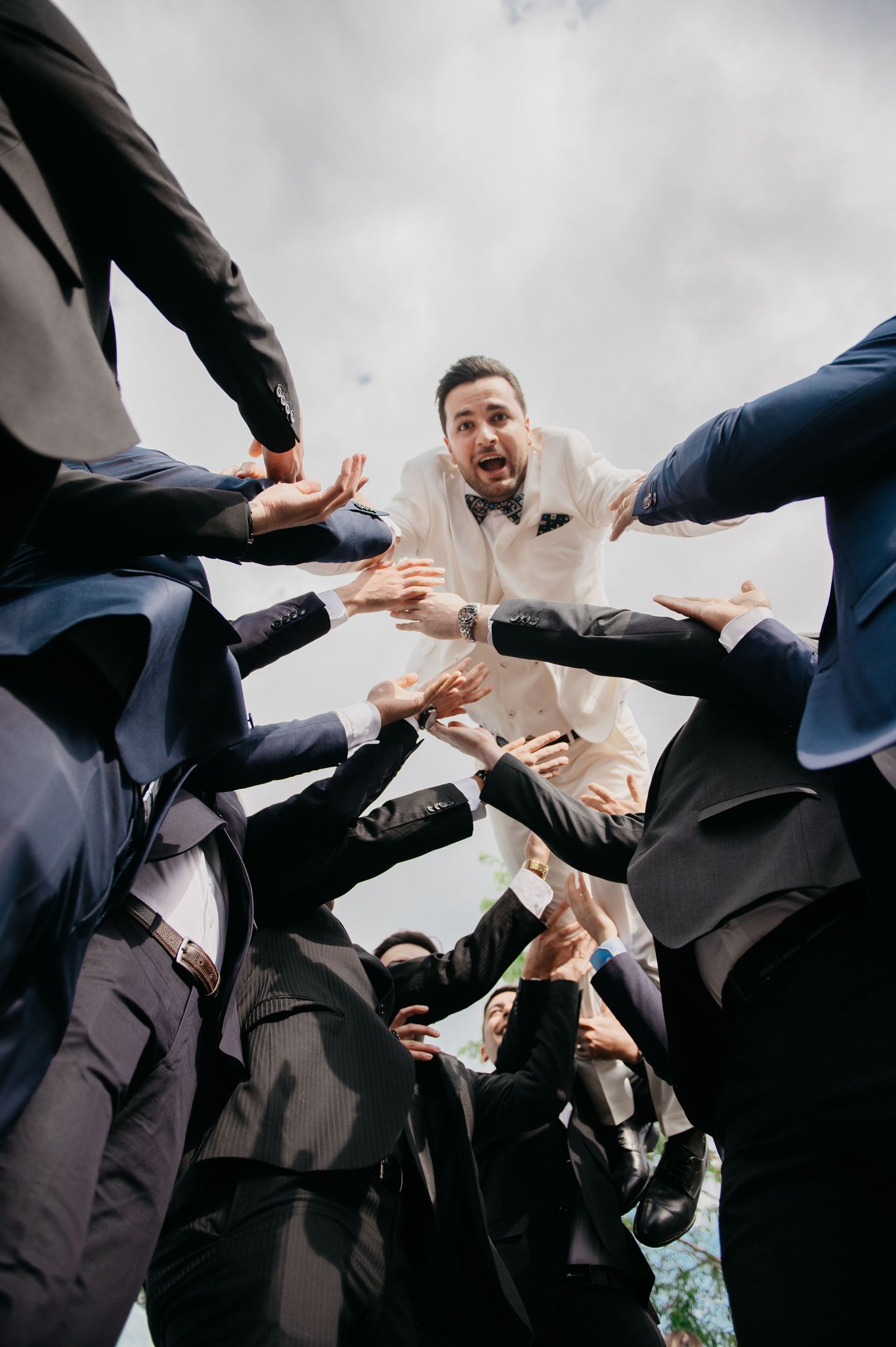 Groom in white tuxedo surrounded by groomsmen in navy suits reaching inward, photographed from below at a wedding.