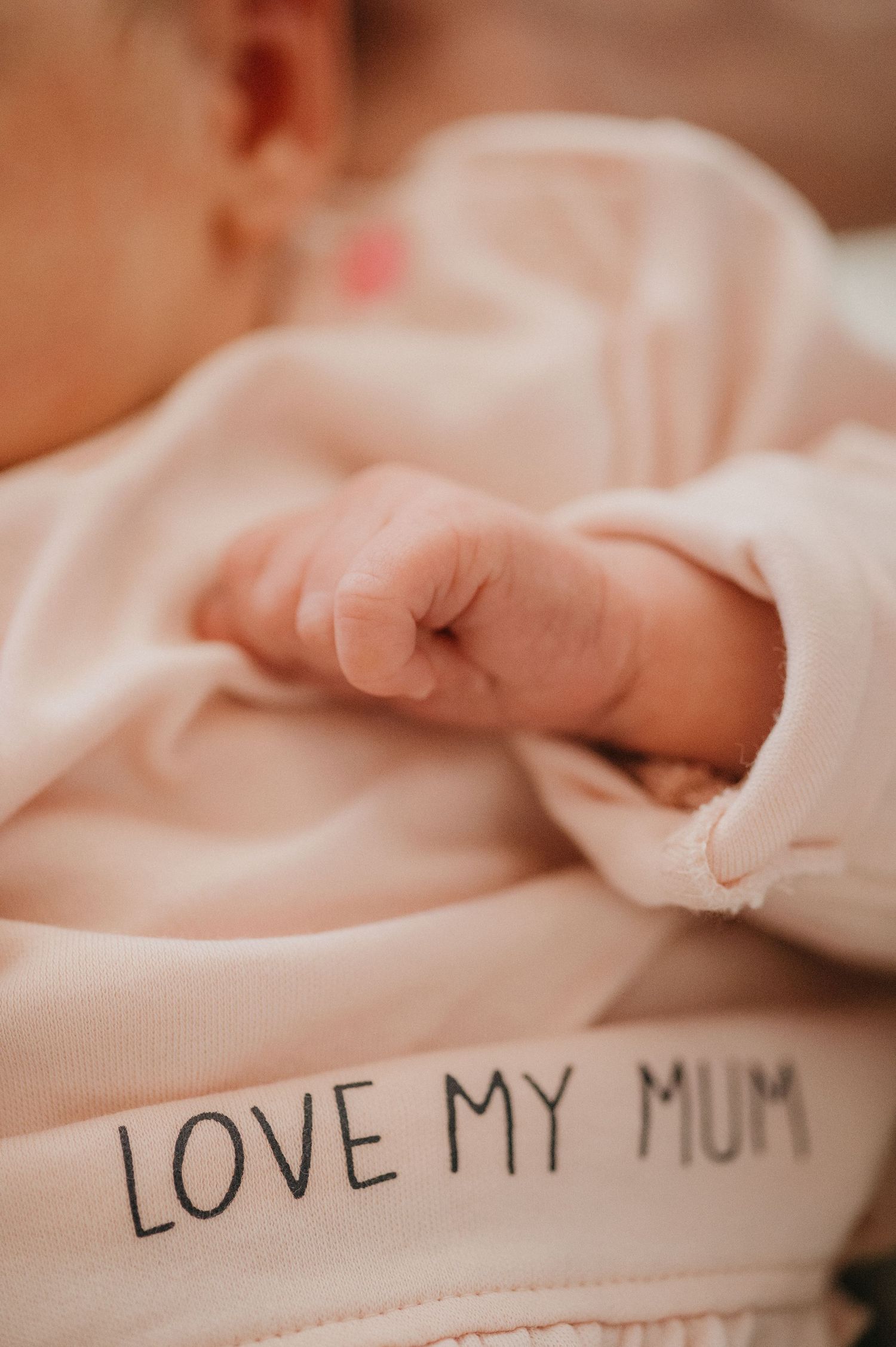 Close-up of a newborn baby's tiny fist resting on a pink onesie that reads Love My Mum.