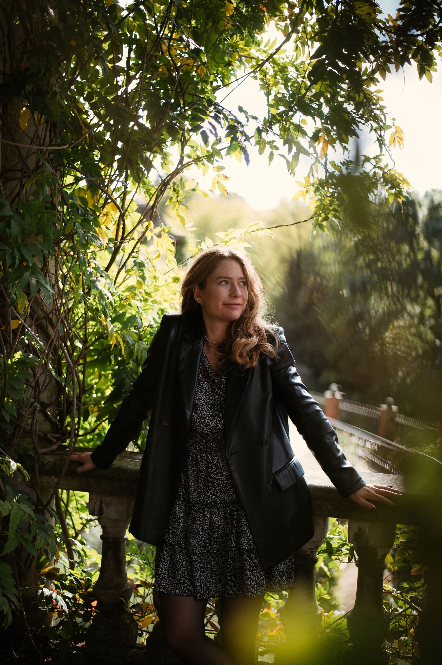 Young woman in leather jacket and floral dress standing by stone railing surrounded by lush green foliage in sunlight.