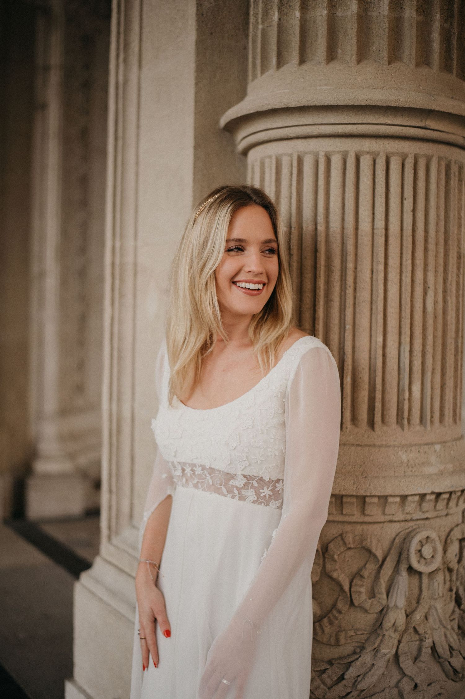 Smiling blonde woman in a white lace wedding gown posing beside ornate stone columns.