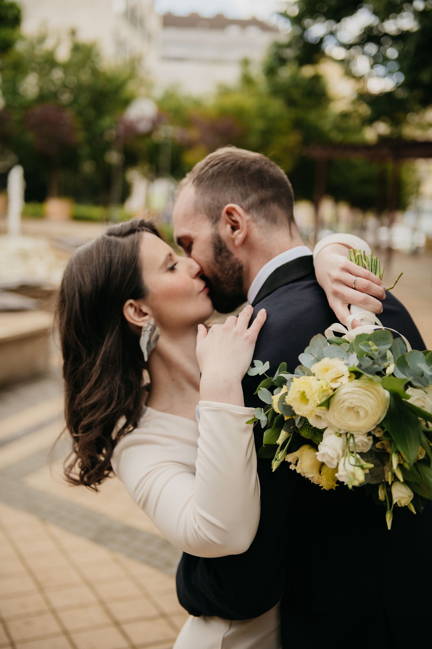 A bride and groom share a romantic kiss outdoors, with the bride holding a bouquet of white and yellow flowers.