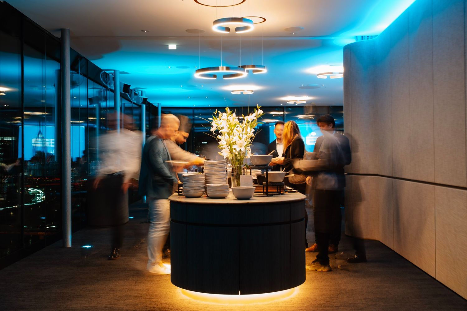 People mingling around a glowing circular bar with floral centerpiece under modern ring lights in a stylish venue.