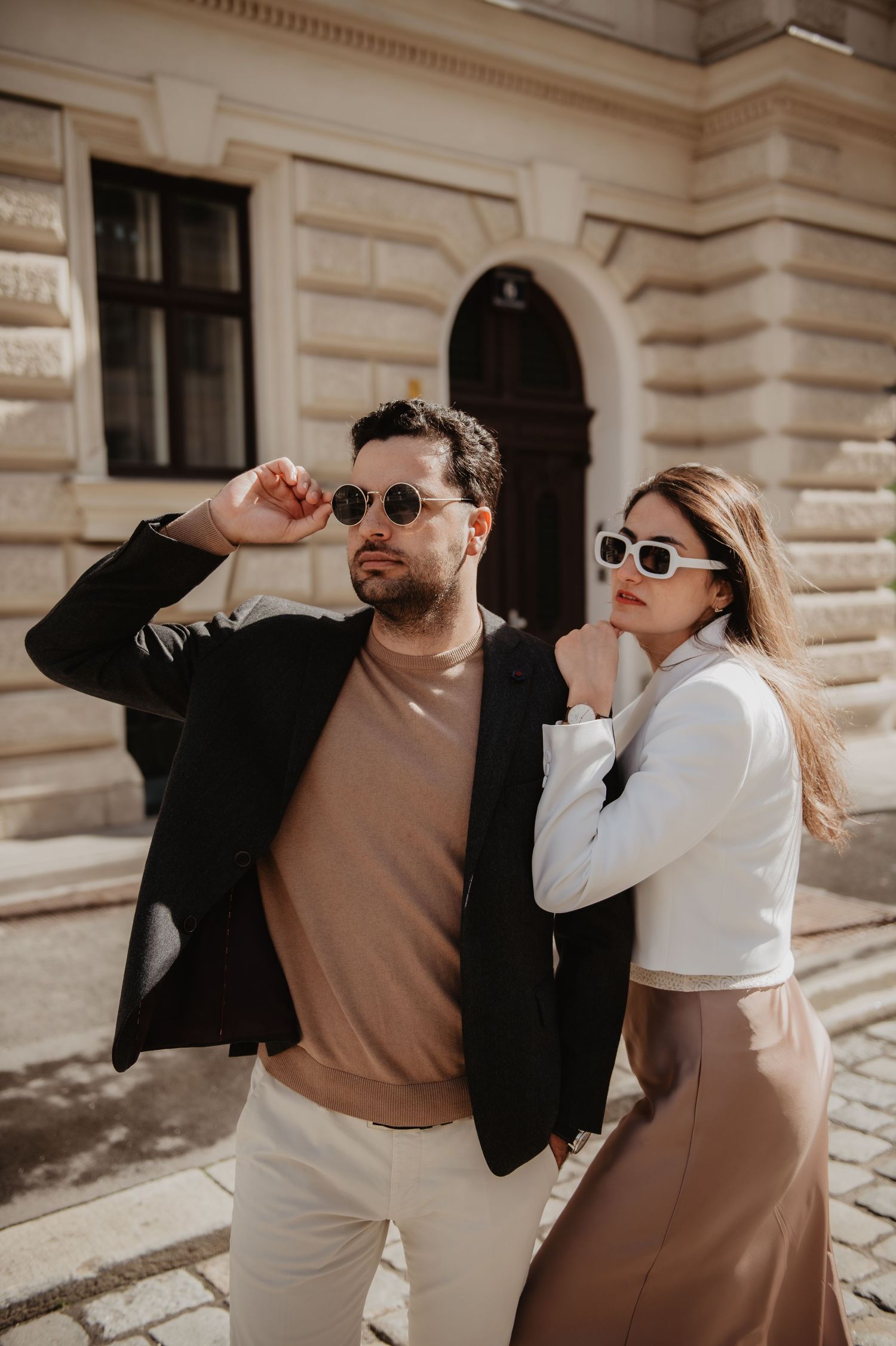 Stylish couple posing outdoors in sunglasses, man in black blazer, woman in white blouse and tan skirt.
