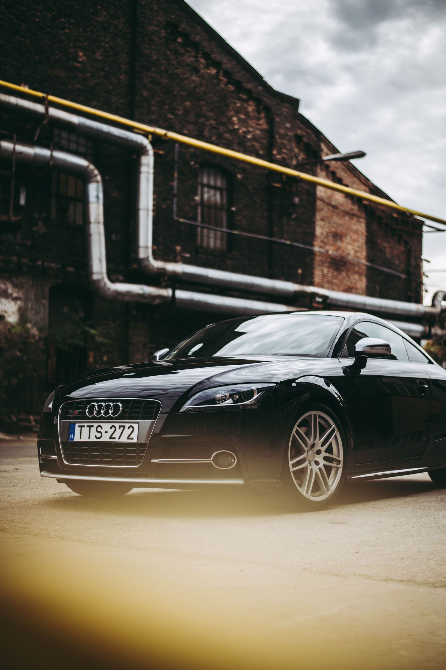 Black Audi TT coupe with license plate TTS-272 parked in front of an industrial brick building with pipes.