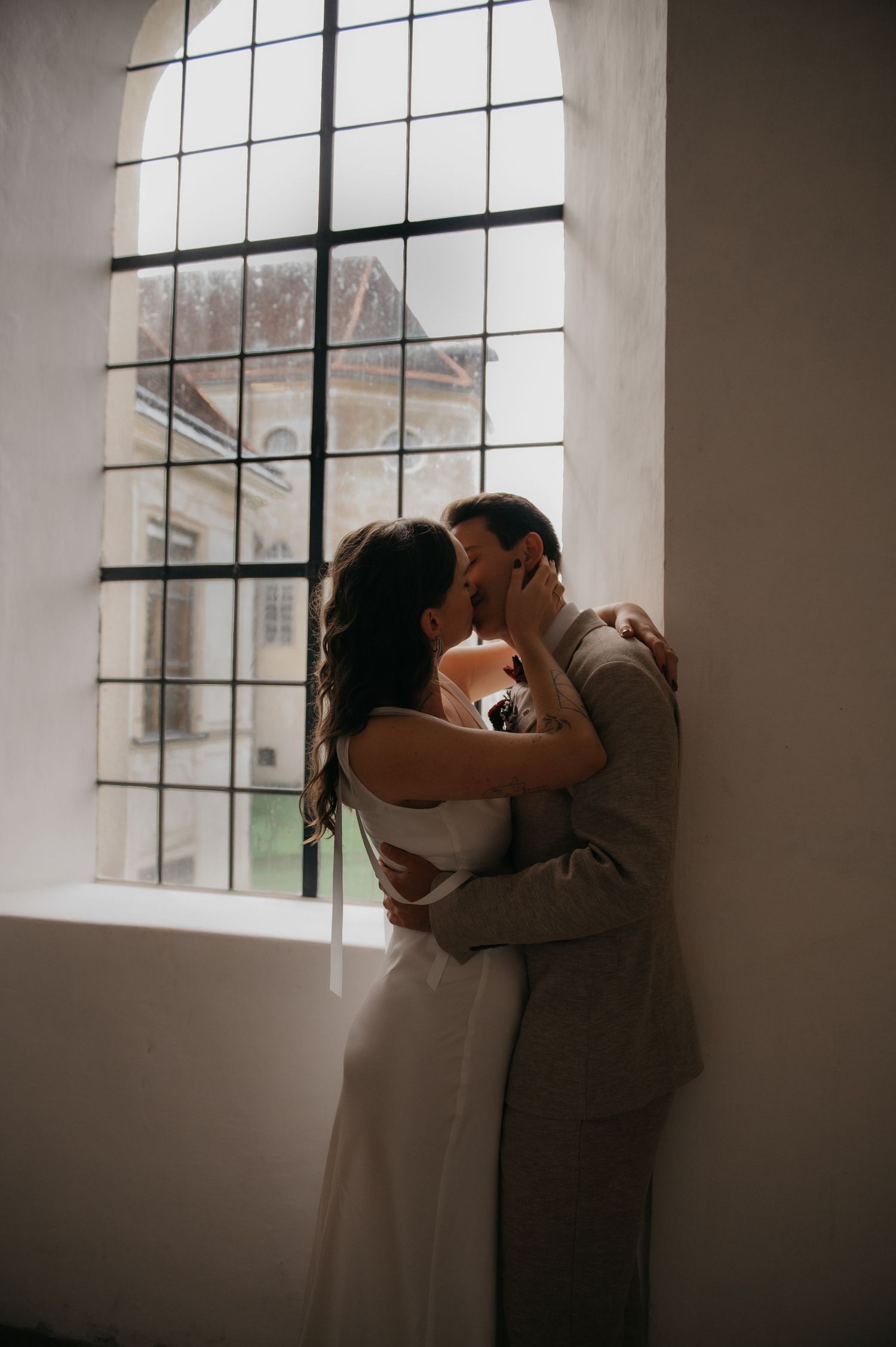 Bride and groom sharing a romantic kiss by a large arched window in a historic venue.