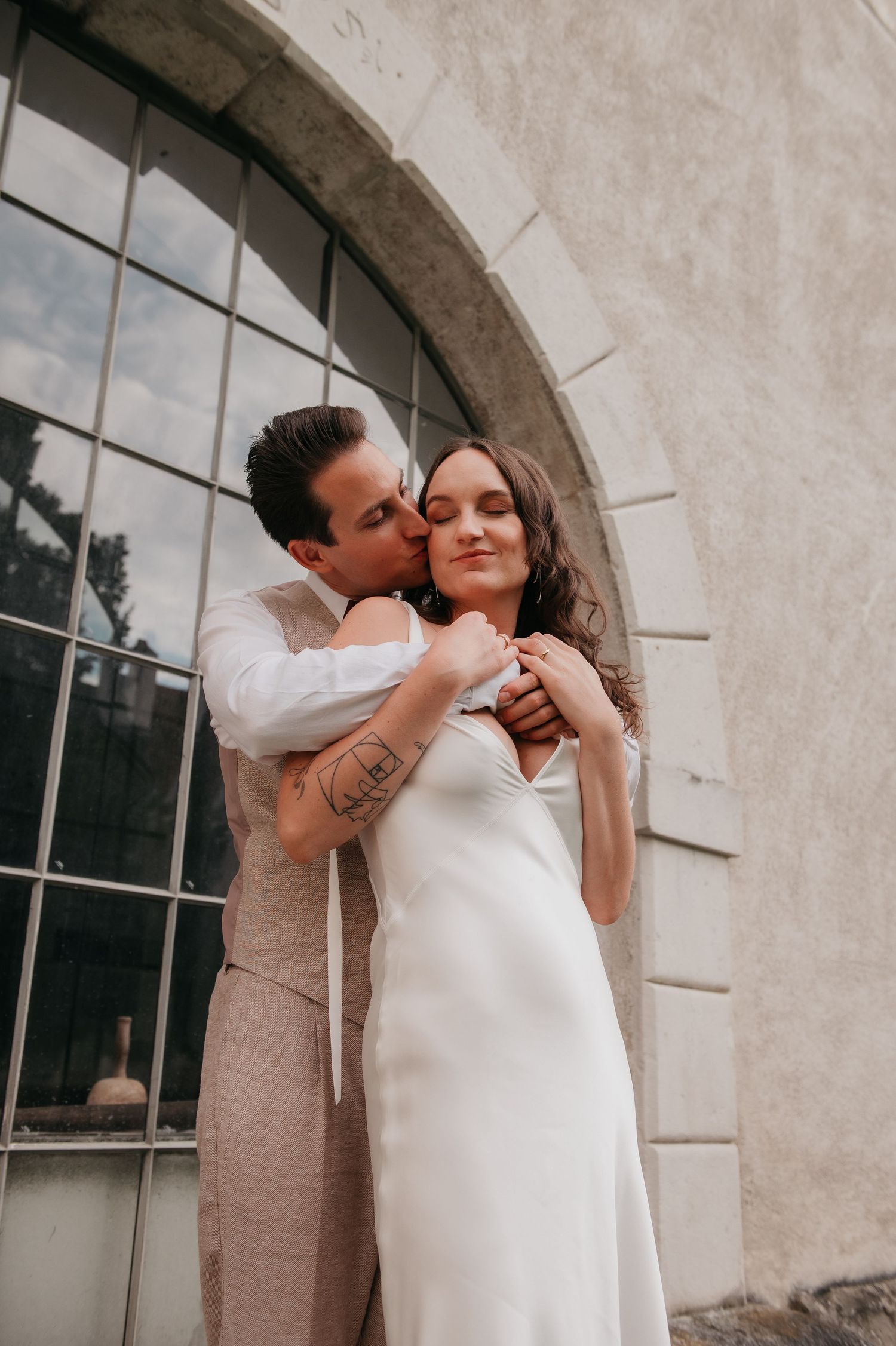 Groom kissing bride on cheek while embracing her in white slip dress outside stone building with arched window.