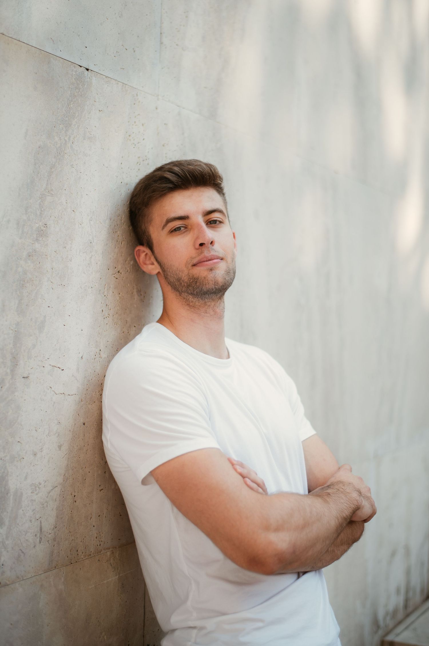 Young man in white t-shirt leaning against concrete wall with arms crossed, confident casual pose.