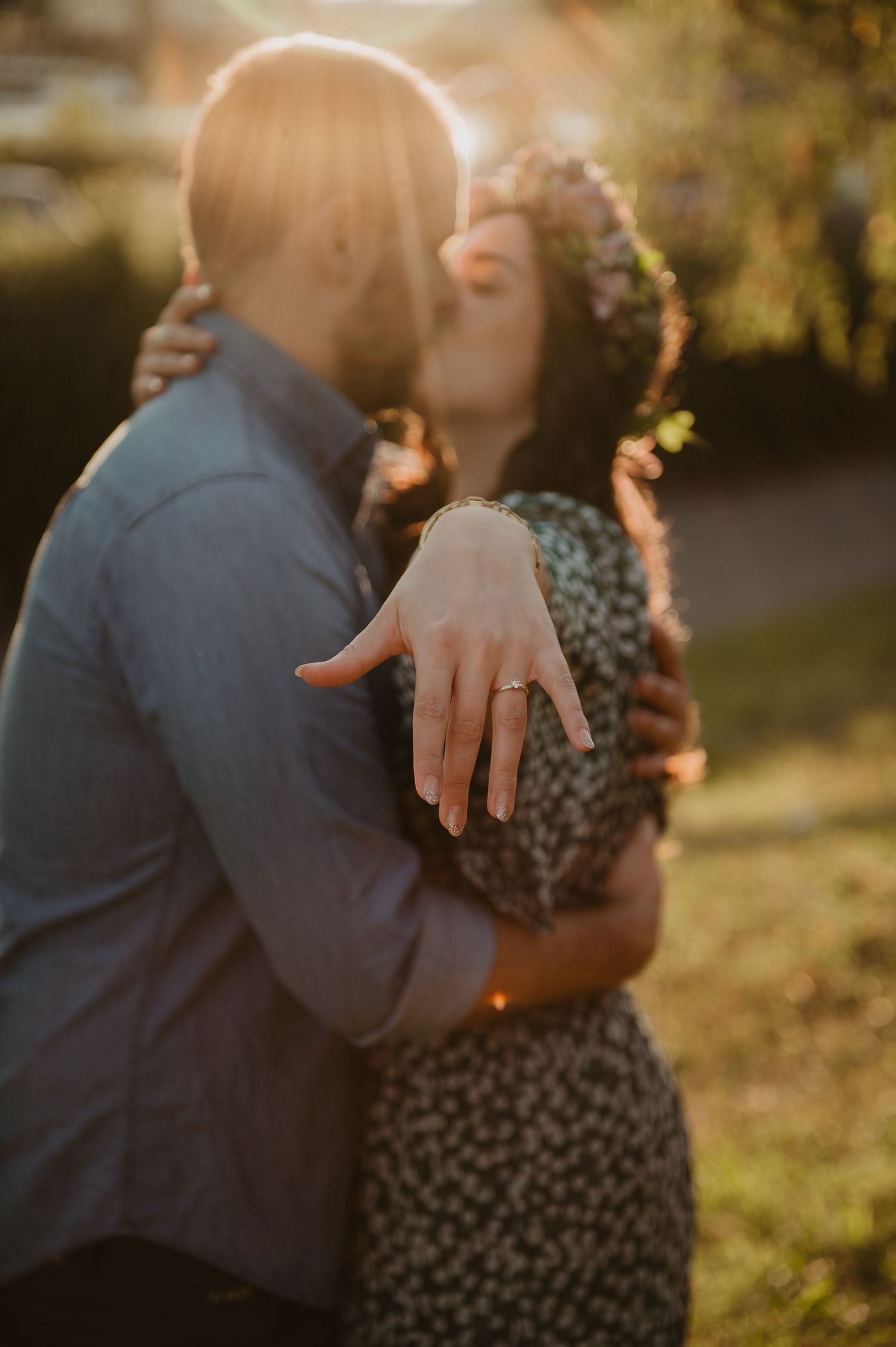 Couple sharing a romantic golden hour kiss outdoors, woman displaying engagement ring while wearing a floral crown.