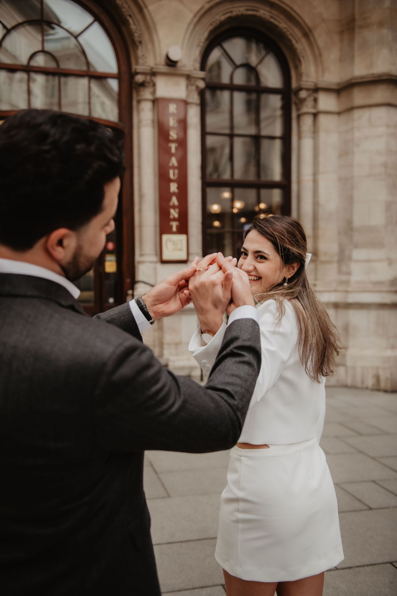 Couple dancing and holding hands in front of a restaurant, woman in white mini dress smiling joyfully.