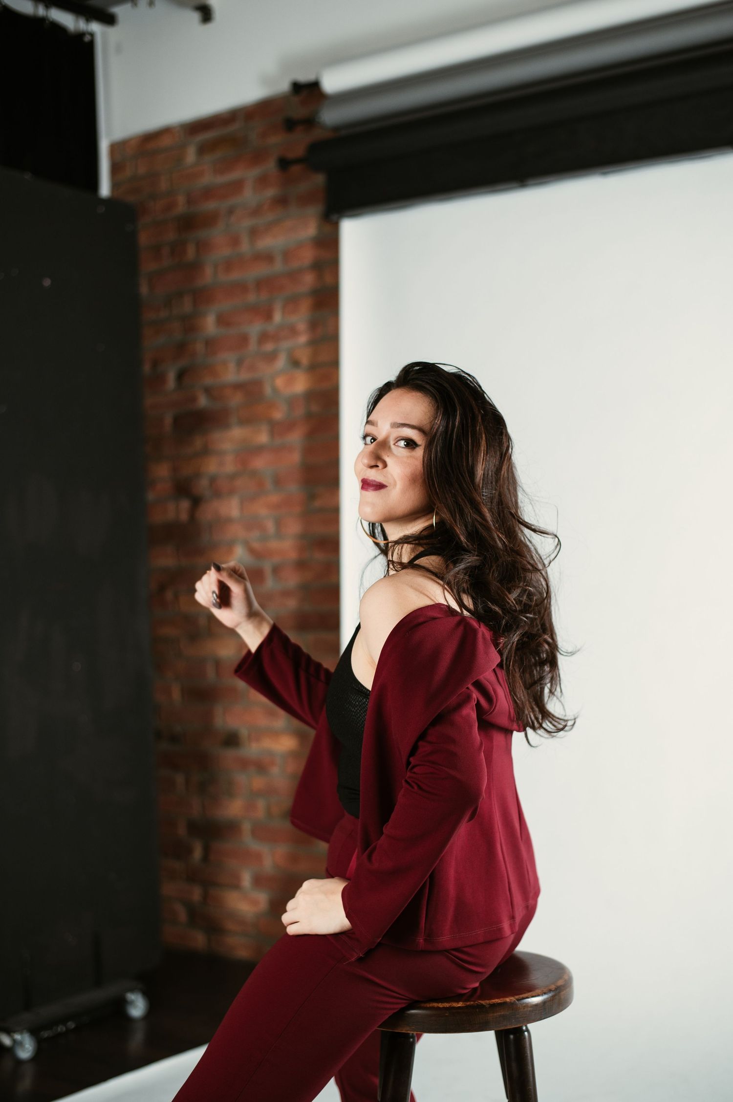 Elegant brunette woman in a burgundy suit sits on a stool in a brick-walled photography studio.
