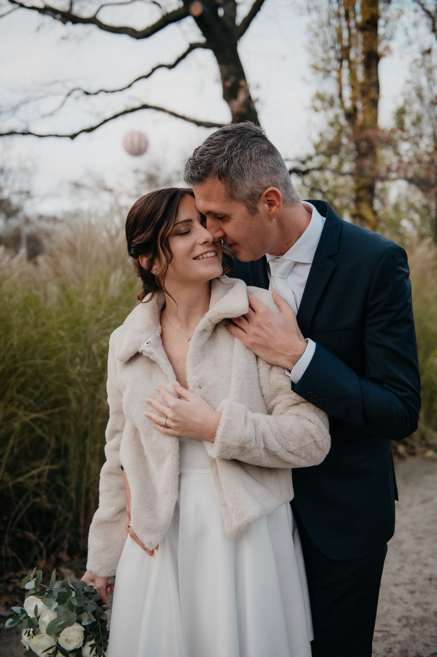 Groom in navy suit embracing smiling bride in fur jacket holding eucalyptus bouquet outdoors in winter.