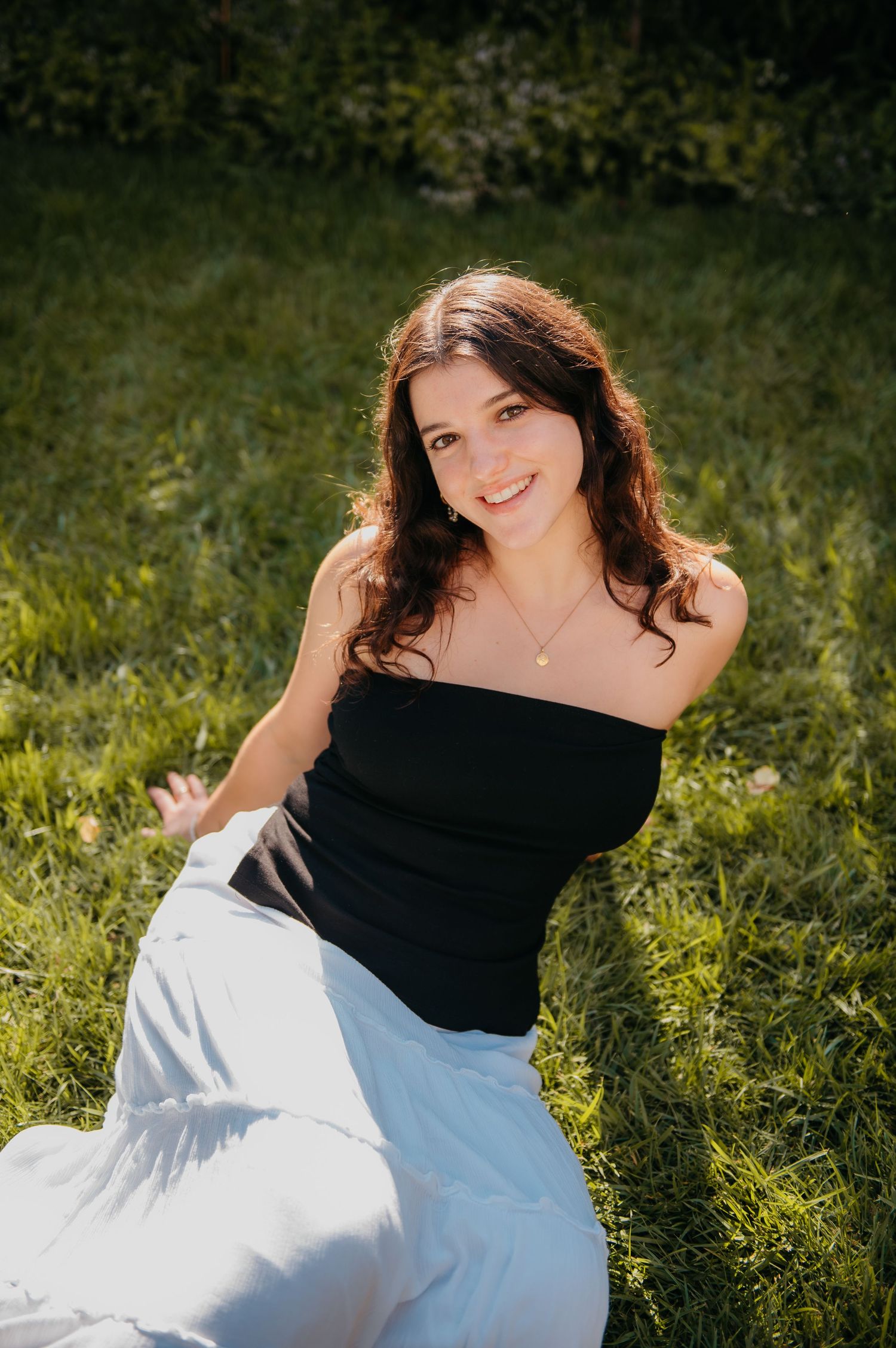 Young woman in black tube top and white skirt sitting on green grass, smiling outdoors in sunlight.