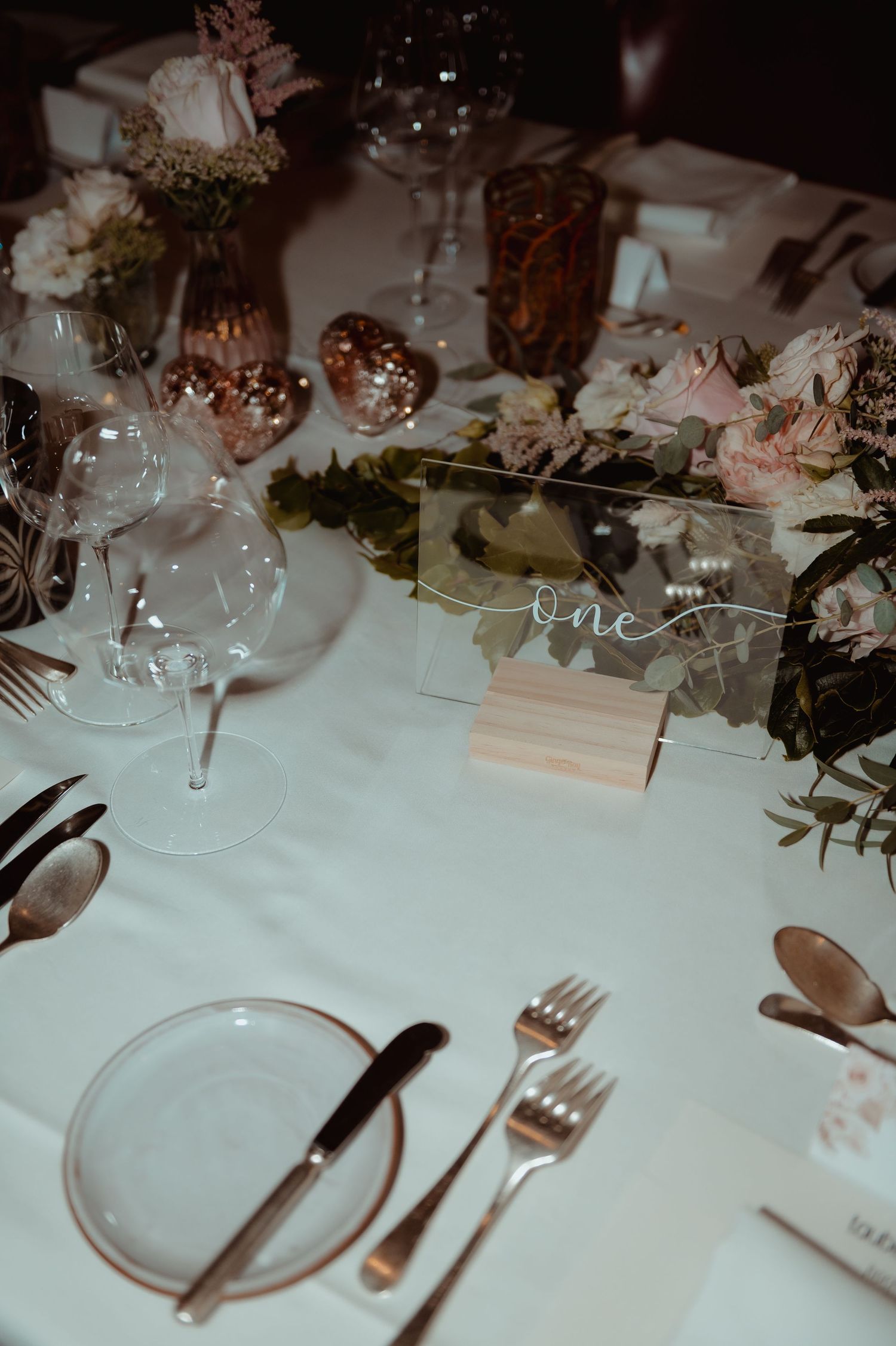 Elegant wedding table setting with acrylic table number, rose gold cutlery, blush floral centerpiece, and glassware on white linen.