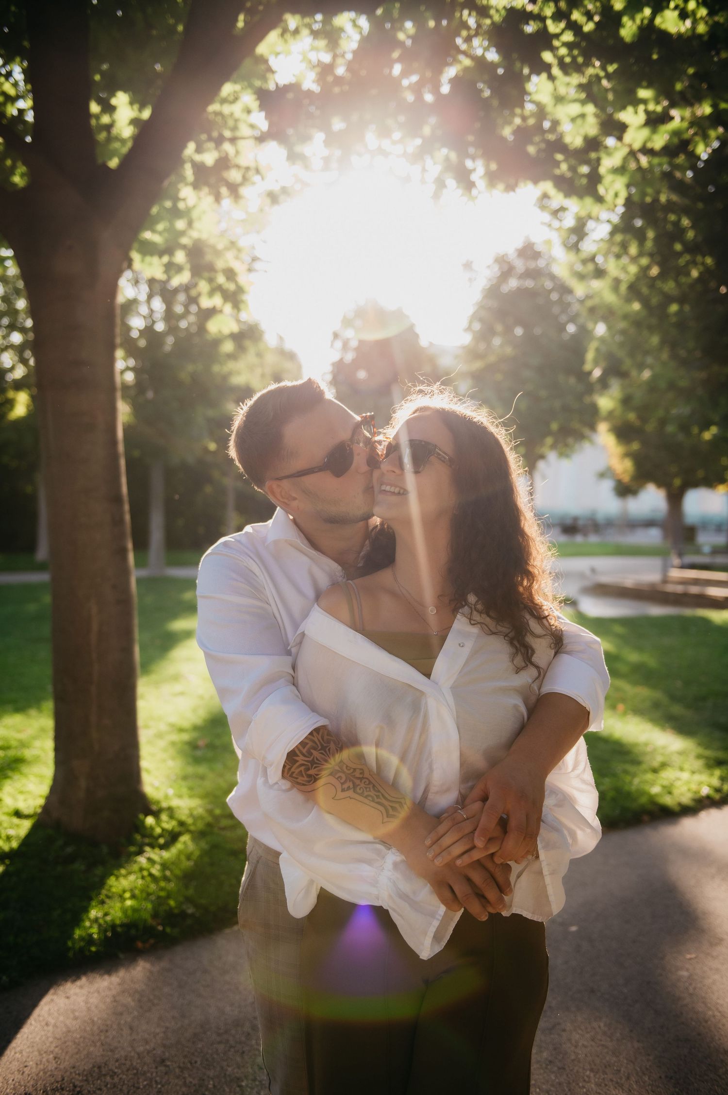 Couple embracing and kissing in sunny park with golden sunburst through green trees backdrop.