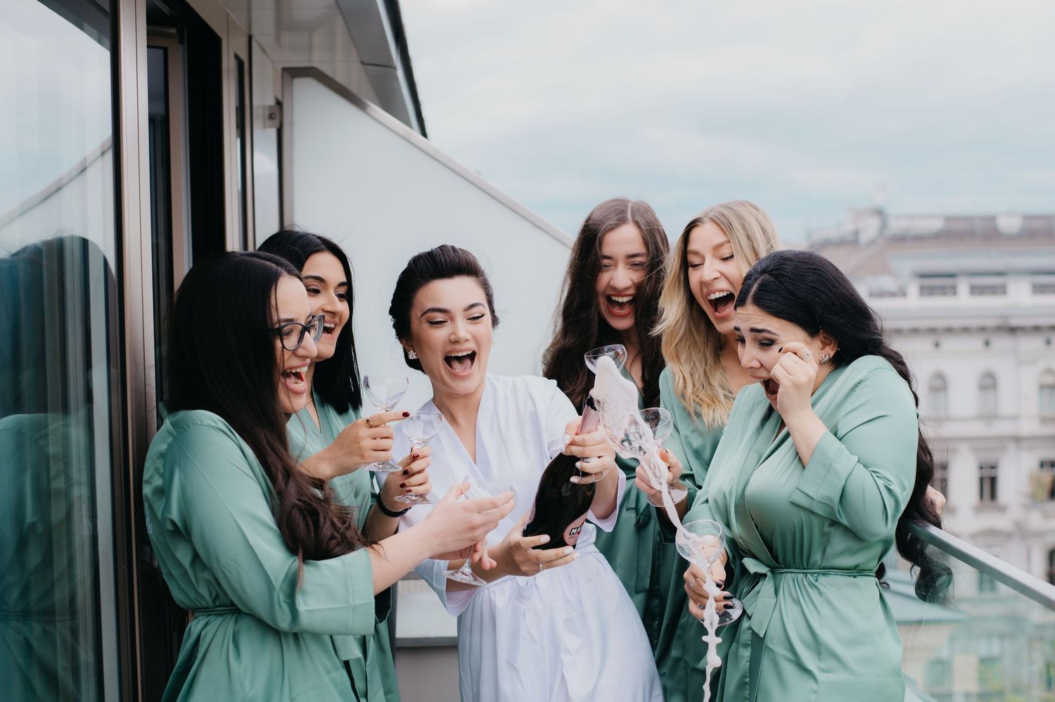 Excited bride in white robe celebrating with bridesmaids in green robes on hotel balcony.