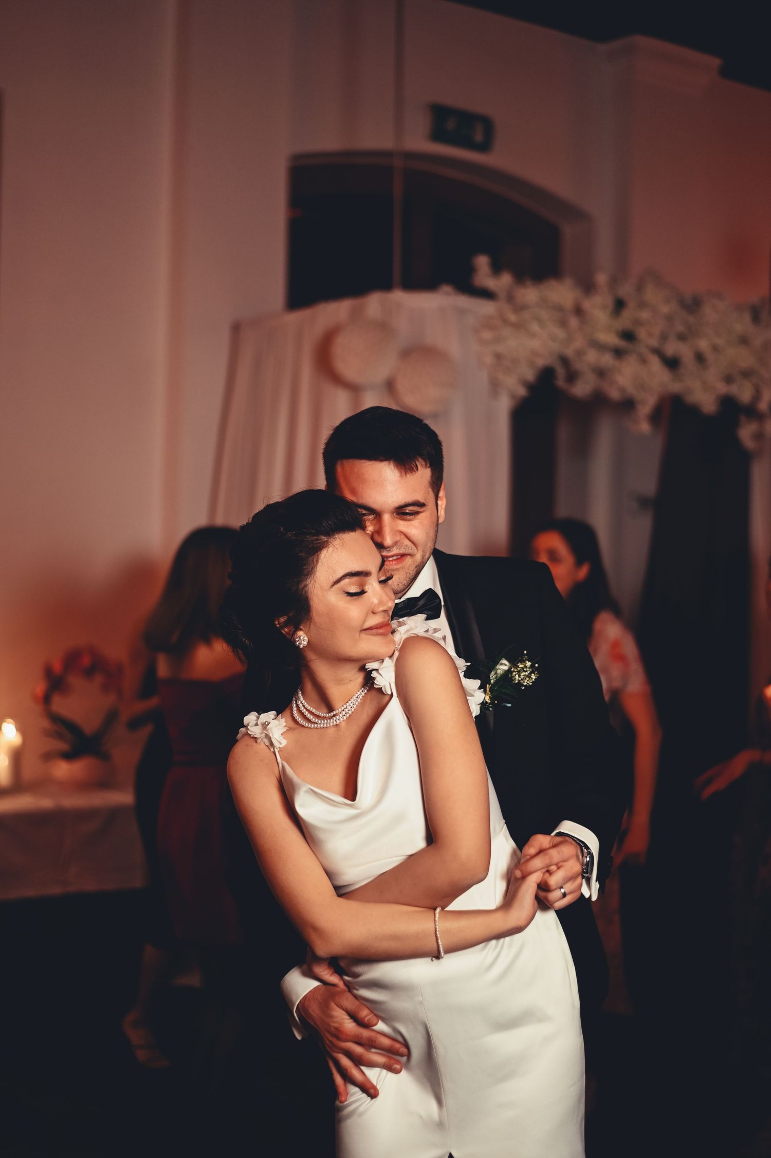 Bride and groom share their first dance, embracing romantically in a warmly lit wedding reception venue.