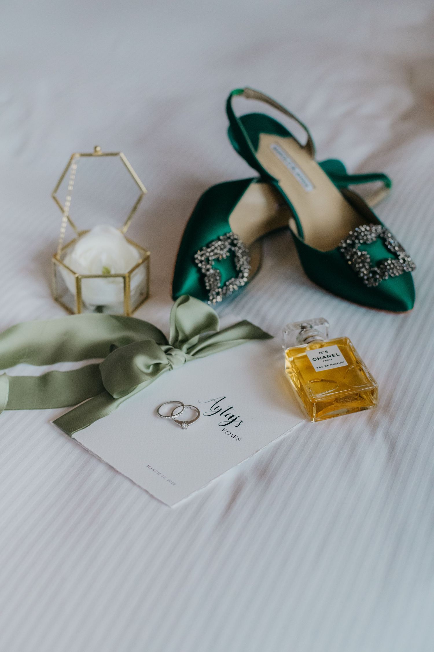 Emerald green Manolo Blahnik heels with crystal buckles beside Chanel No.5 perfume, wedding rings, and sage ribbon.