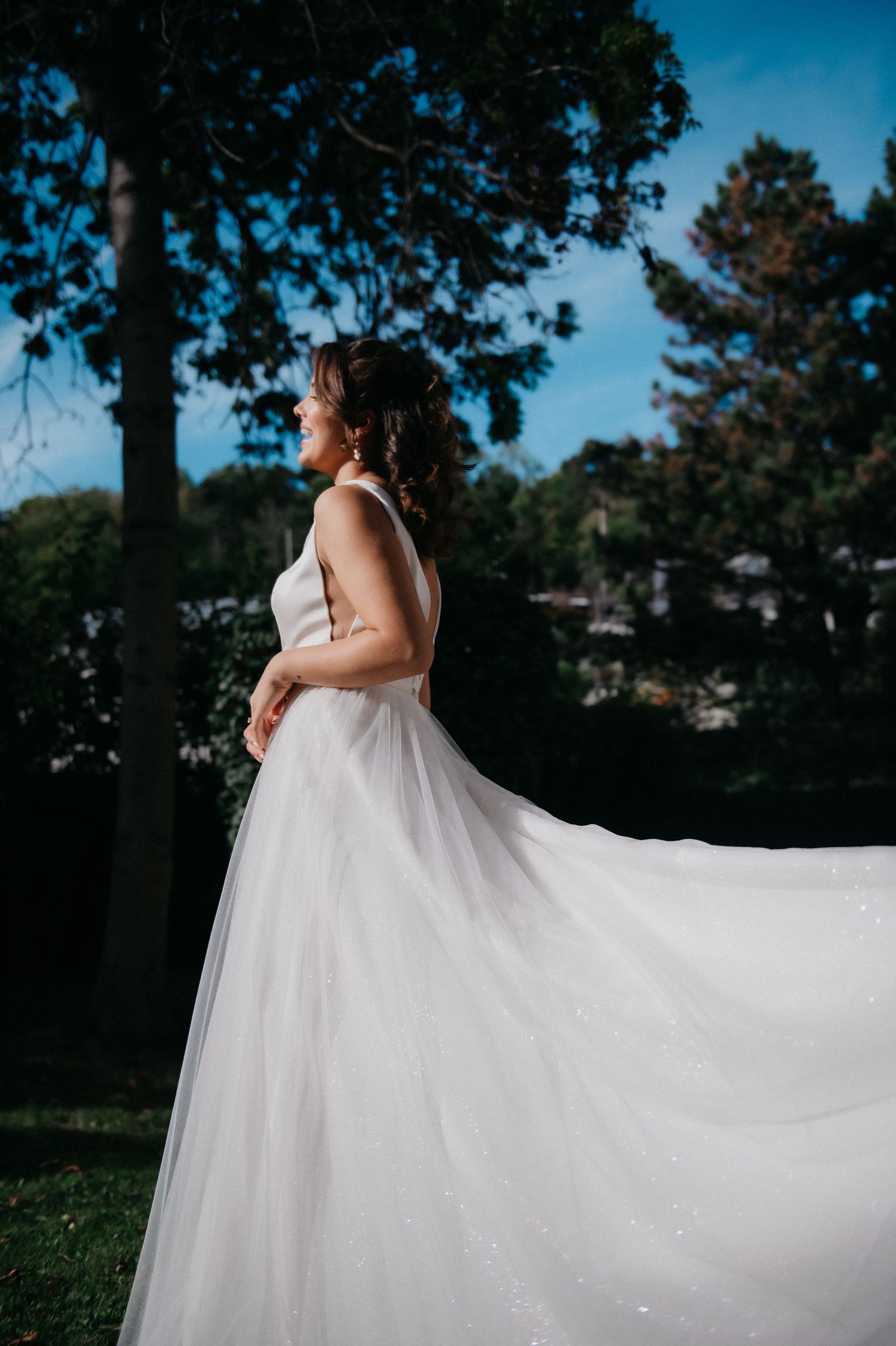 Bride in flowing white tulle wedding gown standing outdoors with trees and blue sky, gown billowing gracefully in the breeze.