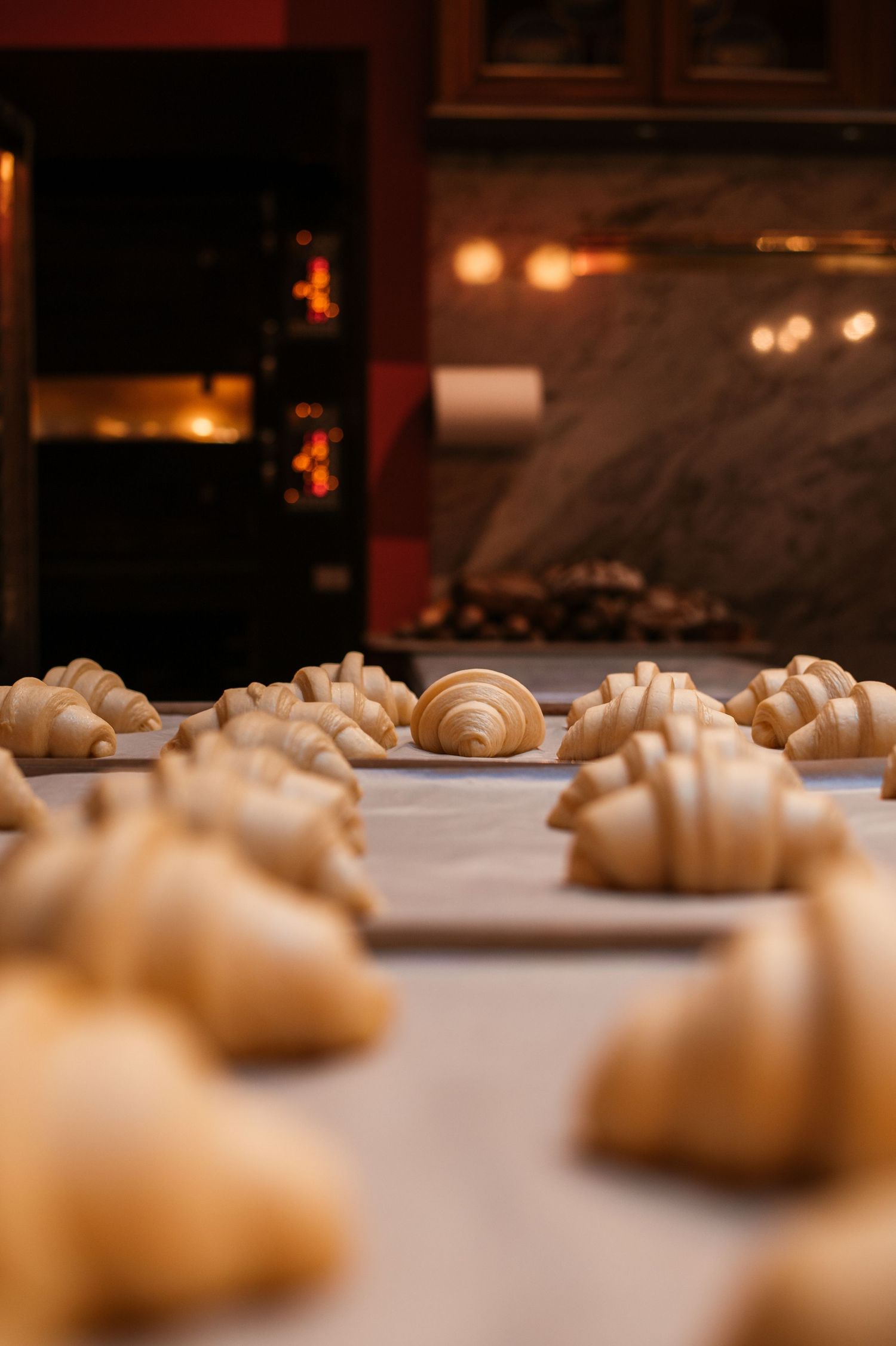 Unbaked croissants arranged on parchment paper in a bakery, with a warm glowing oven in the background.