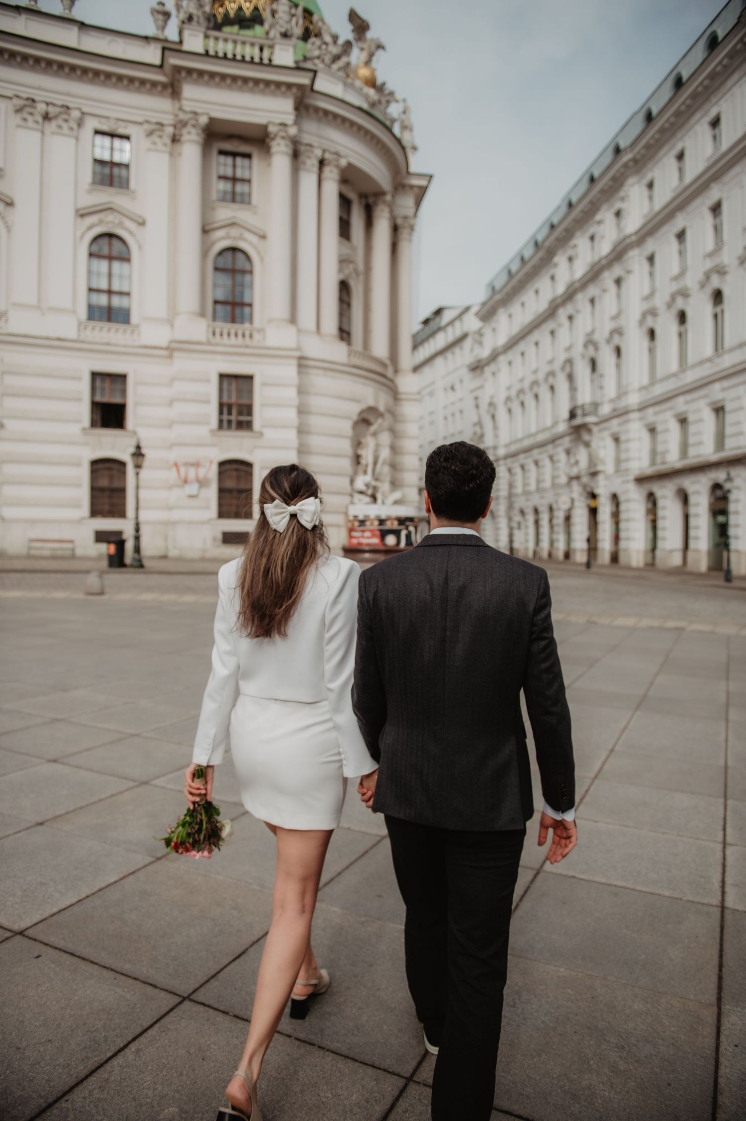 Couple in wedding attire walking hand-in-hand past grand European architecture, bride holding bouquet.