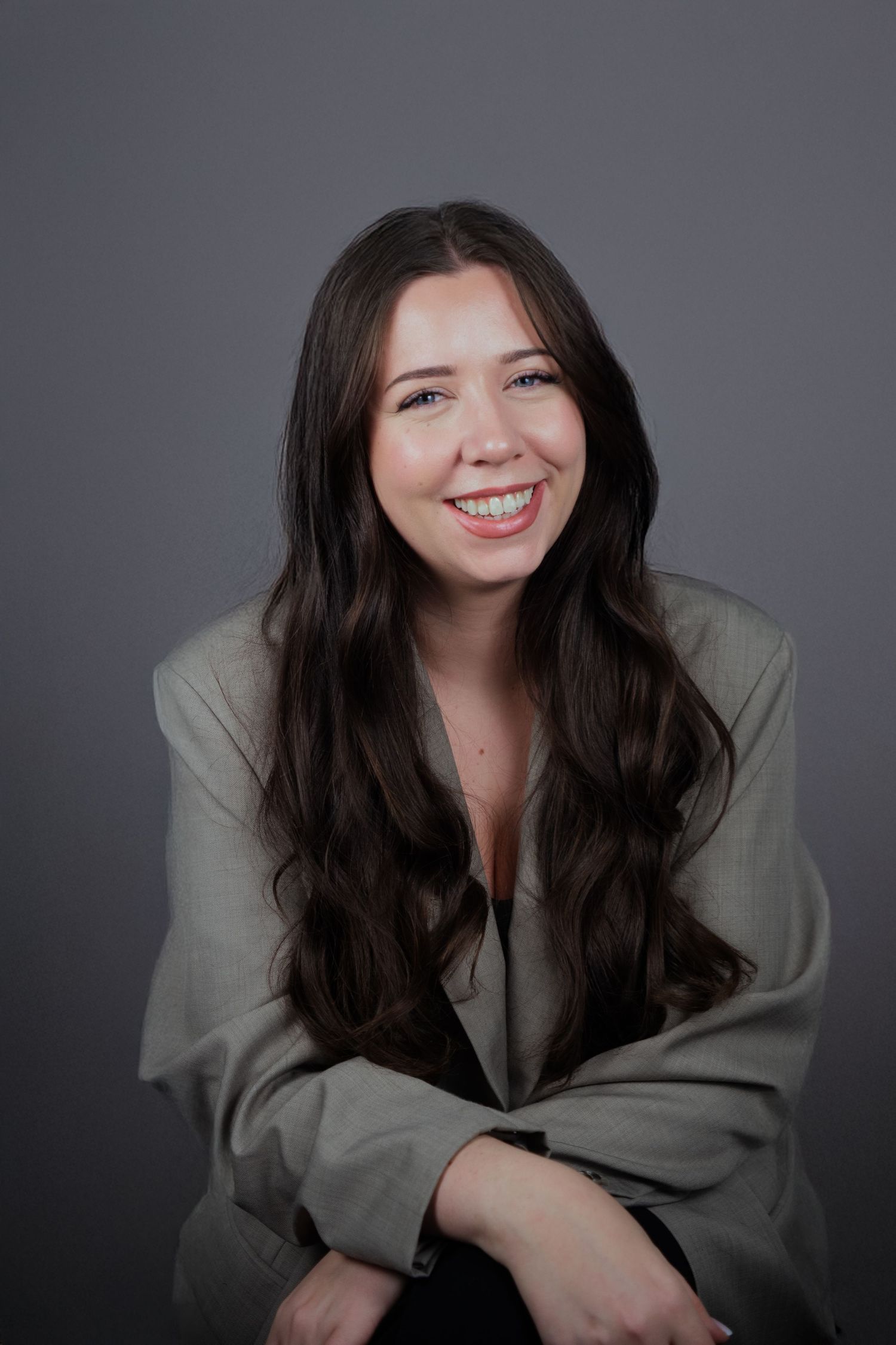 Young woman with long brown hair smiling in a gray blazer, professional headshot on gray background.