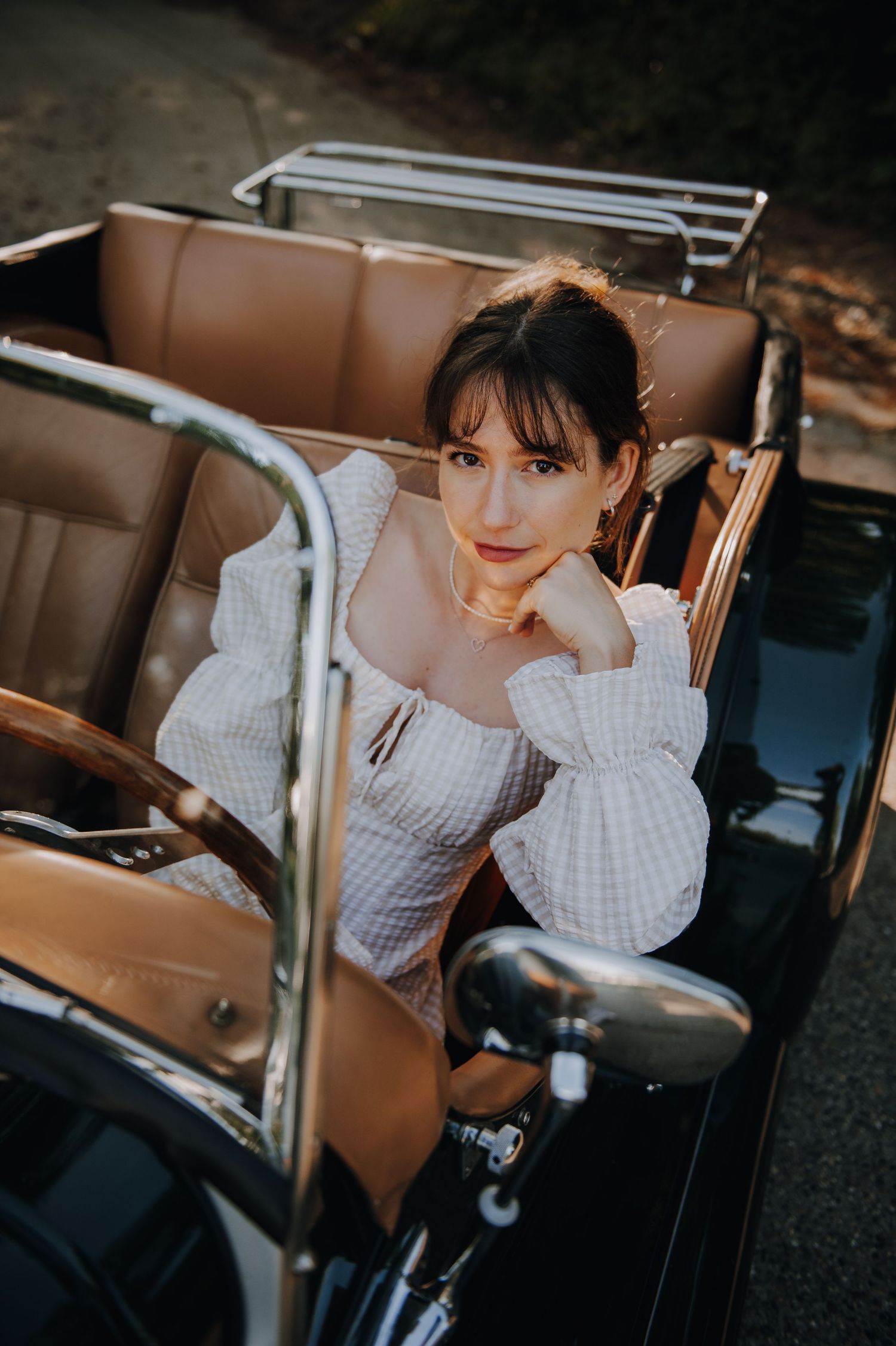 Young woman in white off-shoulder dress poses elegantly in a vintage black convertible car with tan leather interior.