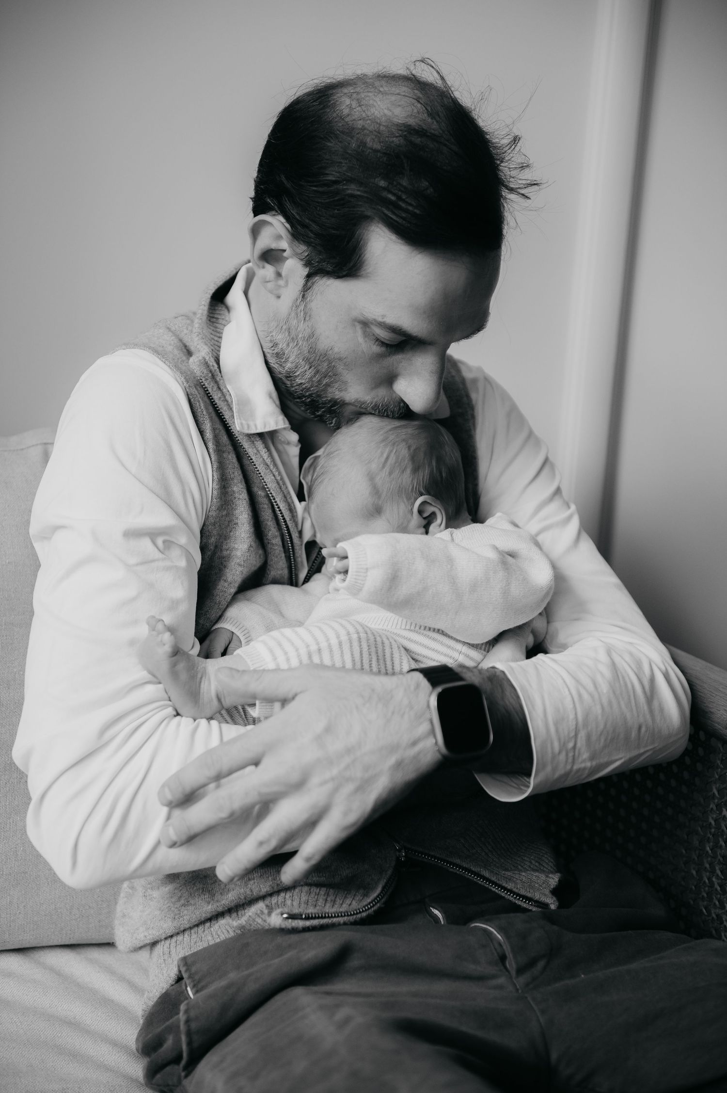 Black and white photo of a bearded father tenderly kissing his newborn baby while cradling it in his arms.