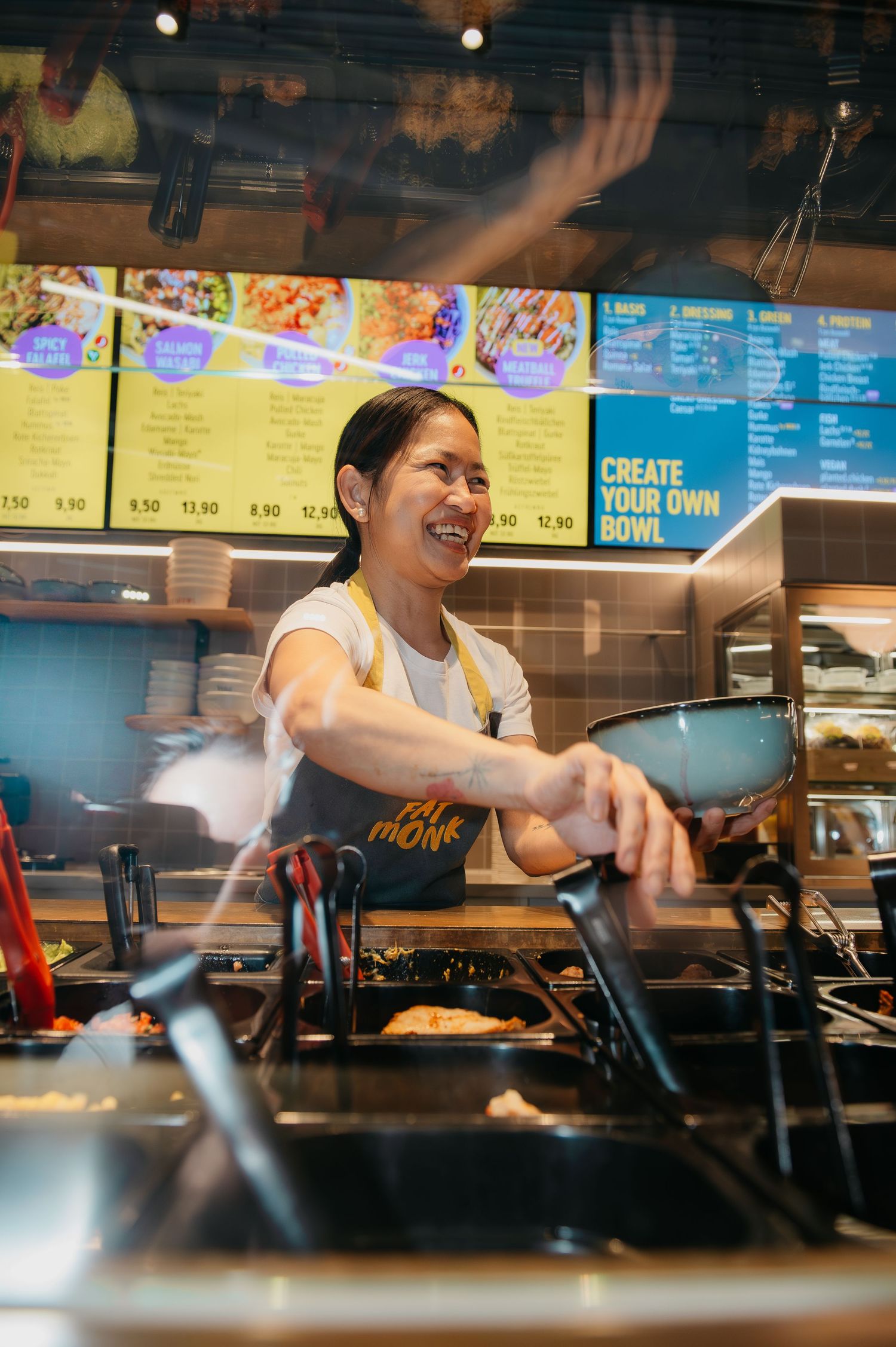 Smiling woman cooking stir-fry in a wok at a fast-casual restaurant with a colorful menu board behind her.