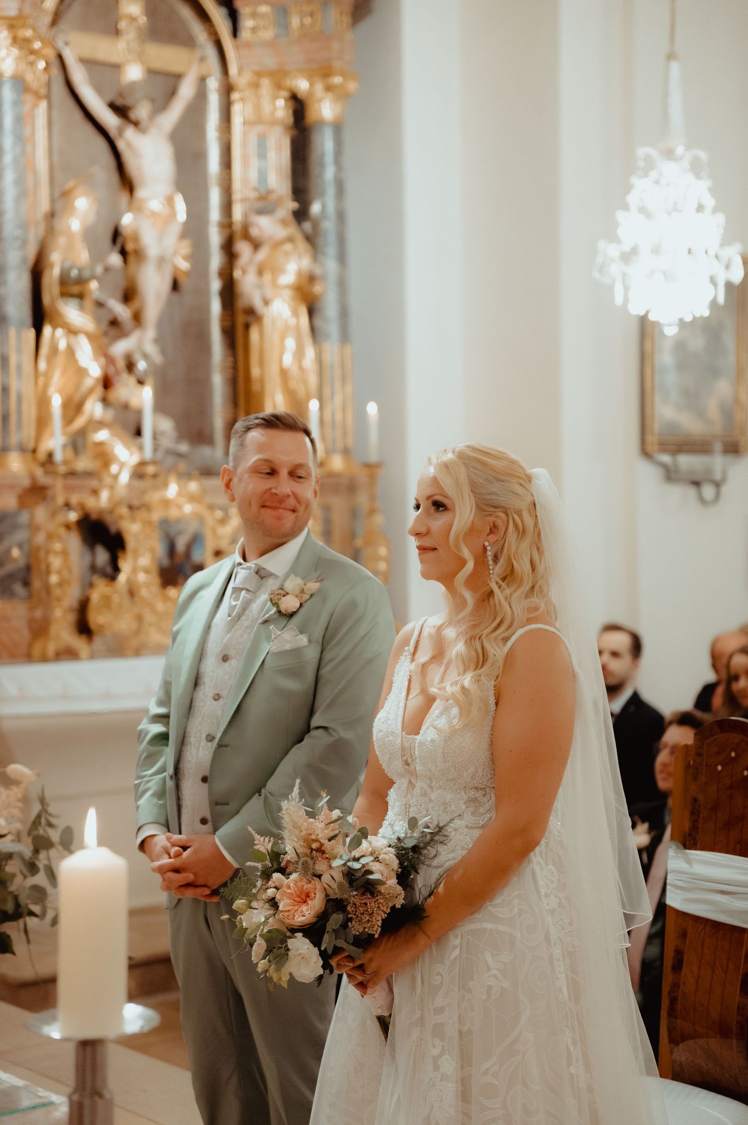 Bride and groom standing at church altar during wedding ceremony, bride holding peach floral bouquet, groom in sage green suit.