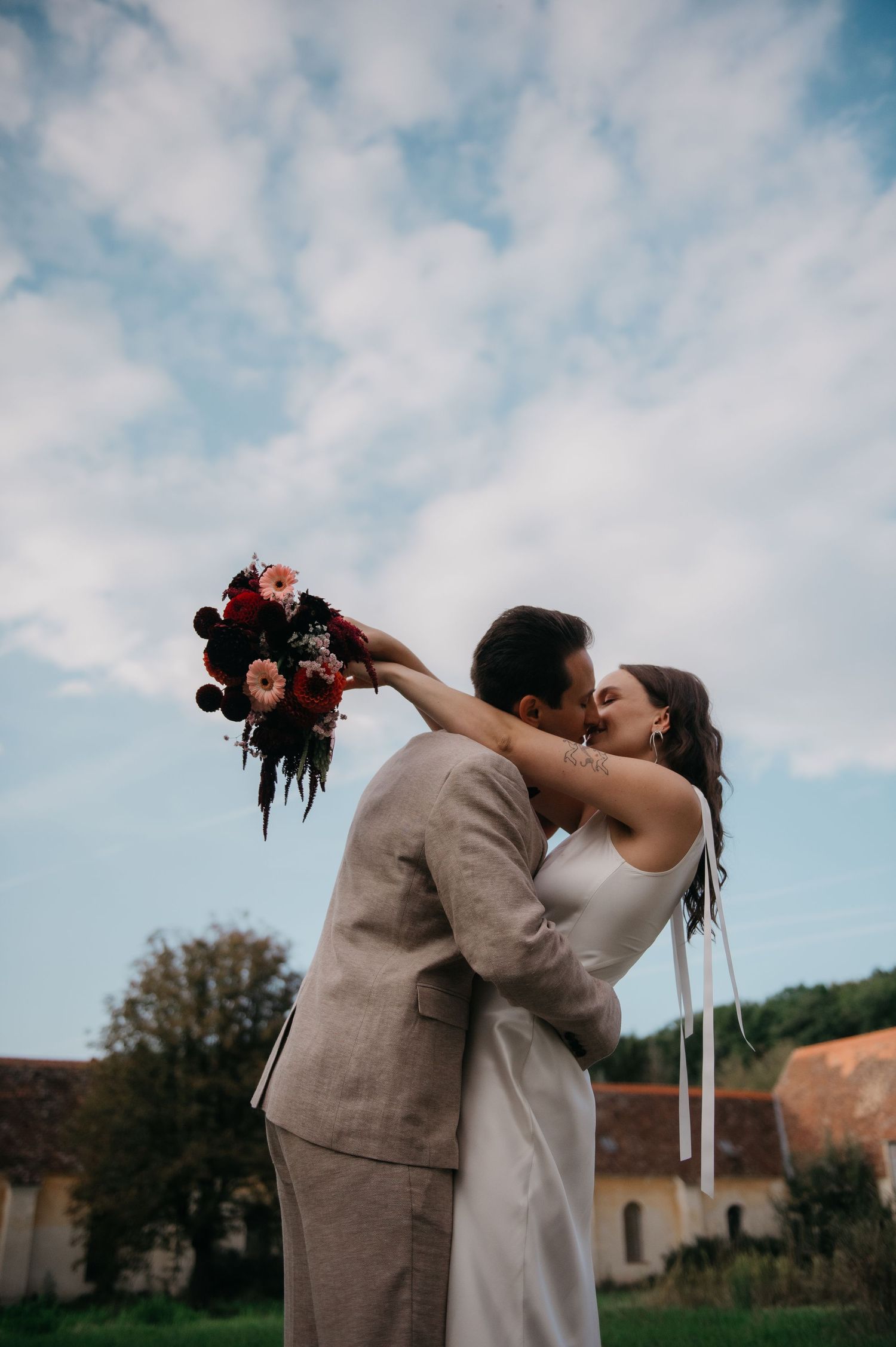 Bride and groom sharing a romantic kiss outdoors, bride holding a vibrant red and pink bouquet against a cloudy sky.