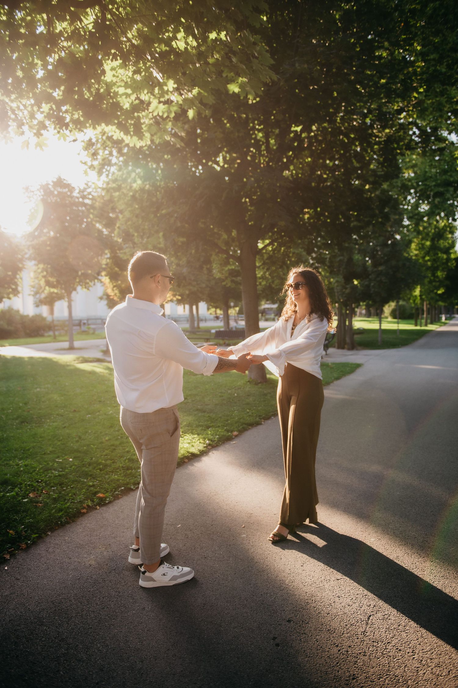 Couple holding hands on a sunlit park path surrounded by lush green trees during golden hour.