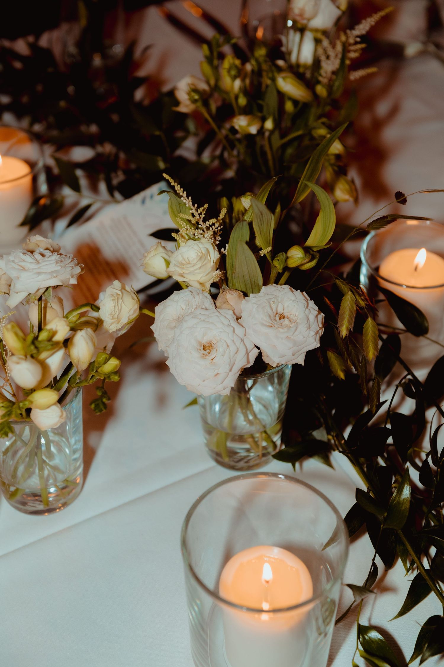 Elegant wedding table centerpiece with white roses in glass vases, greenery, and glowing pillar candles on white linen.
