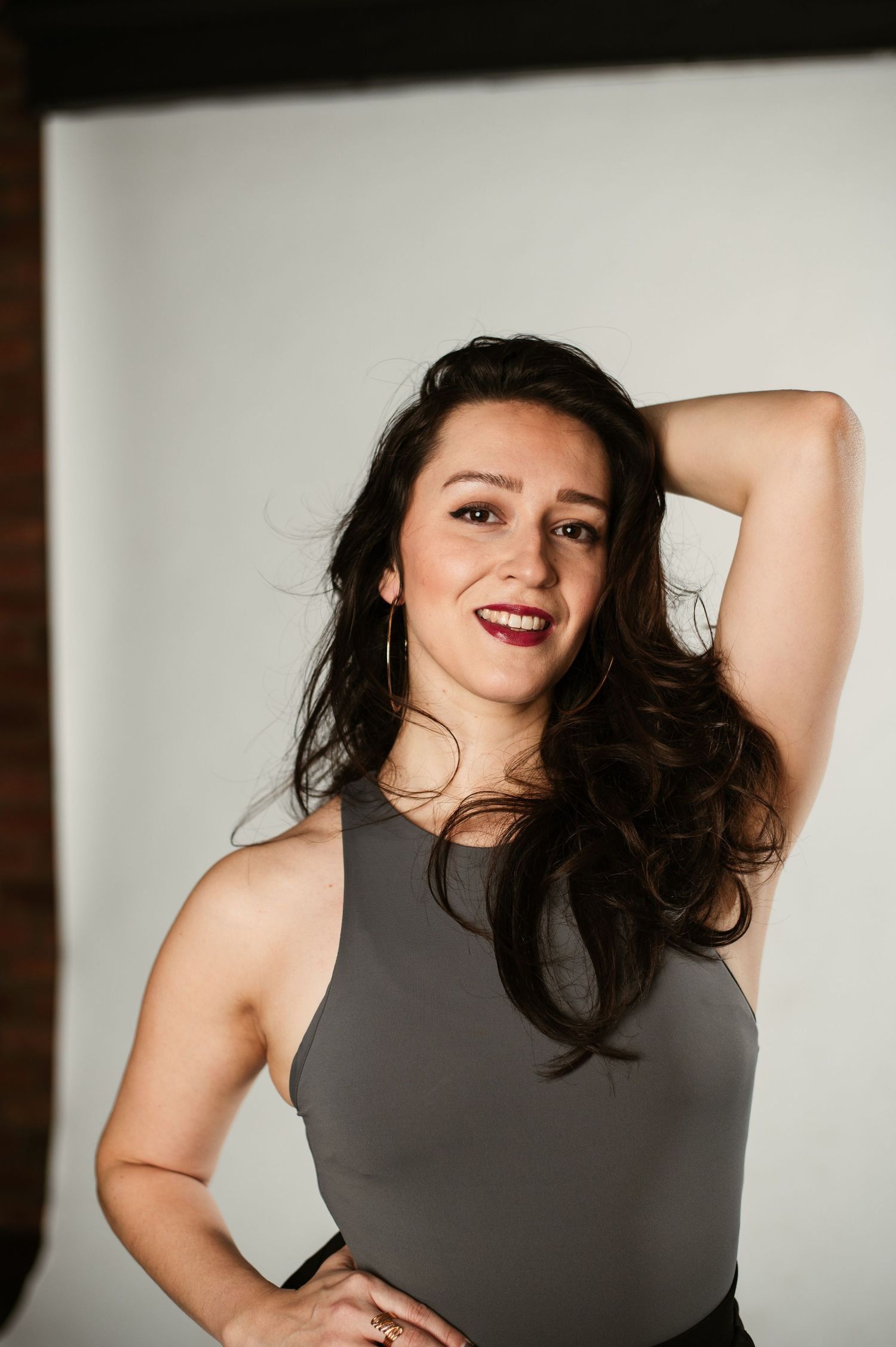 Confident brunette woman in a gray sleeveless bodysuit poses with hand behind head in a studio.
