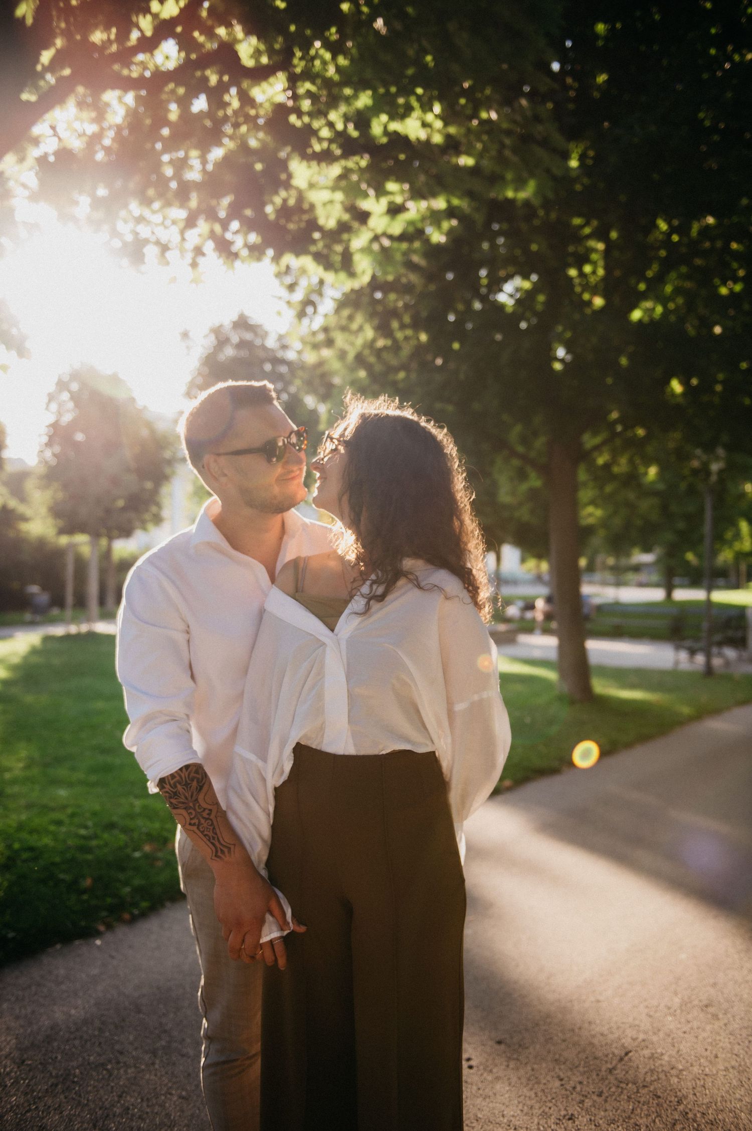 Couple sharing a tender moment on a sunny park pathway bathed in warm golden sunlight among green trees.
