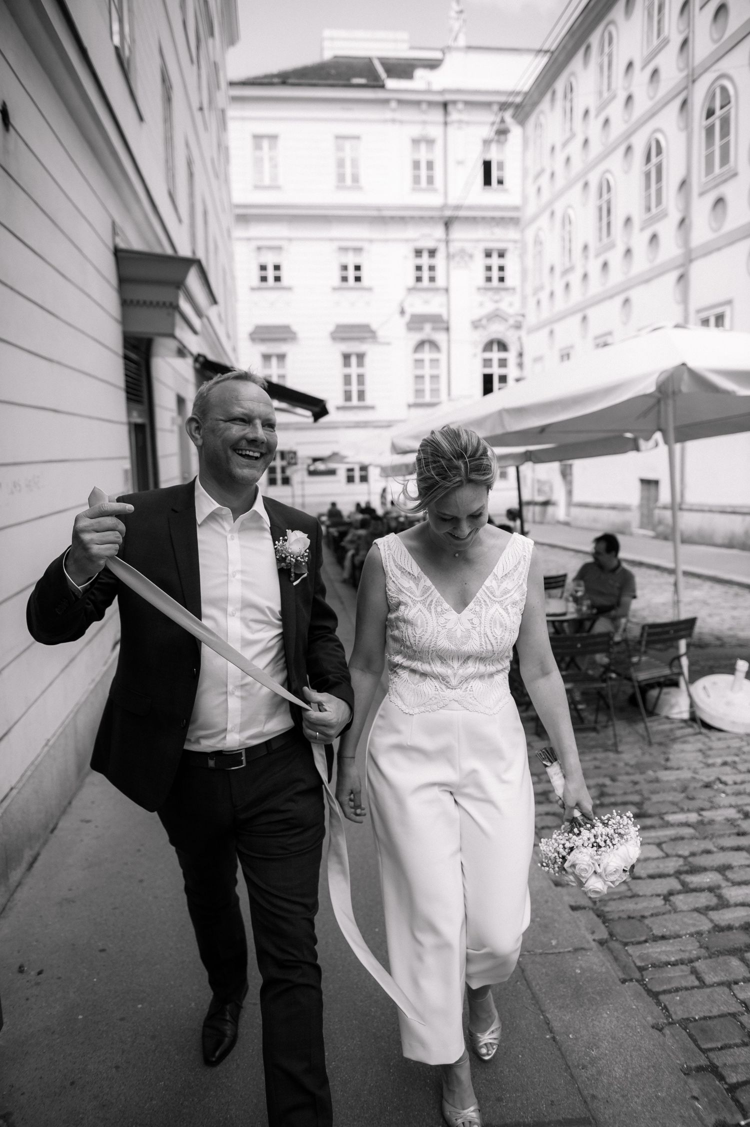Black and white photo of a smiling groom and bride in white jumpsuit walking on a cobblestone street.