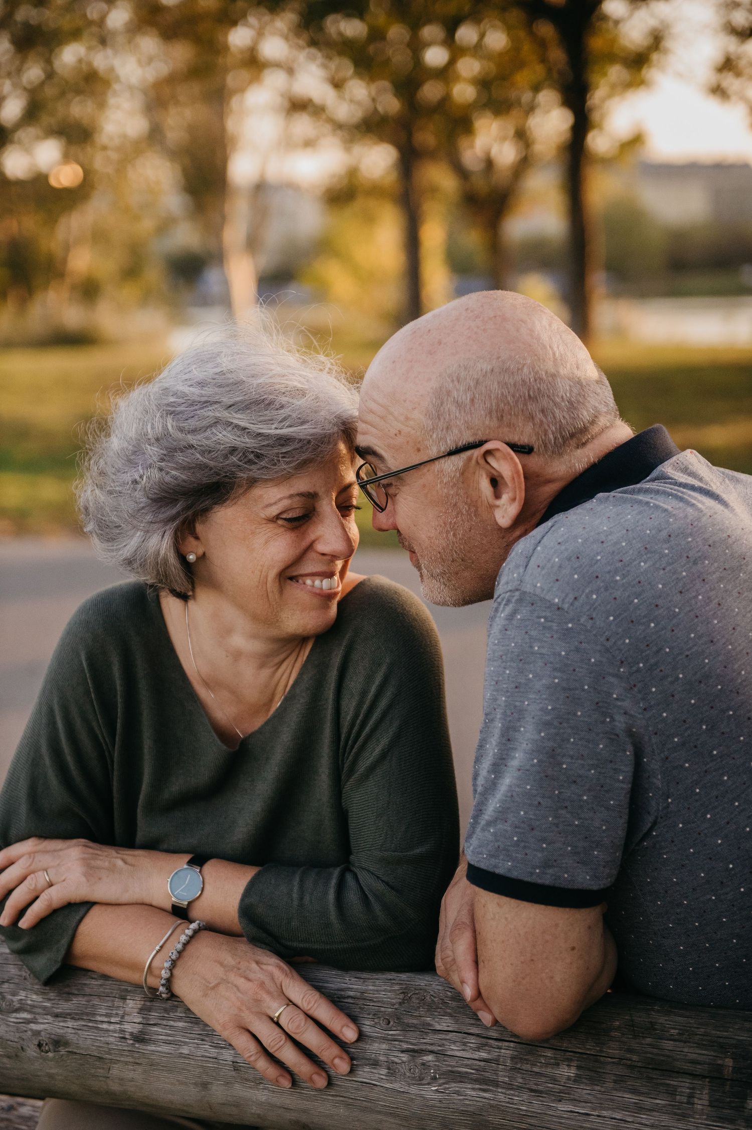 An elderly couple shares a tender moment on a park bench, smiling and touching foreheads in golden hour light.