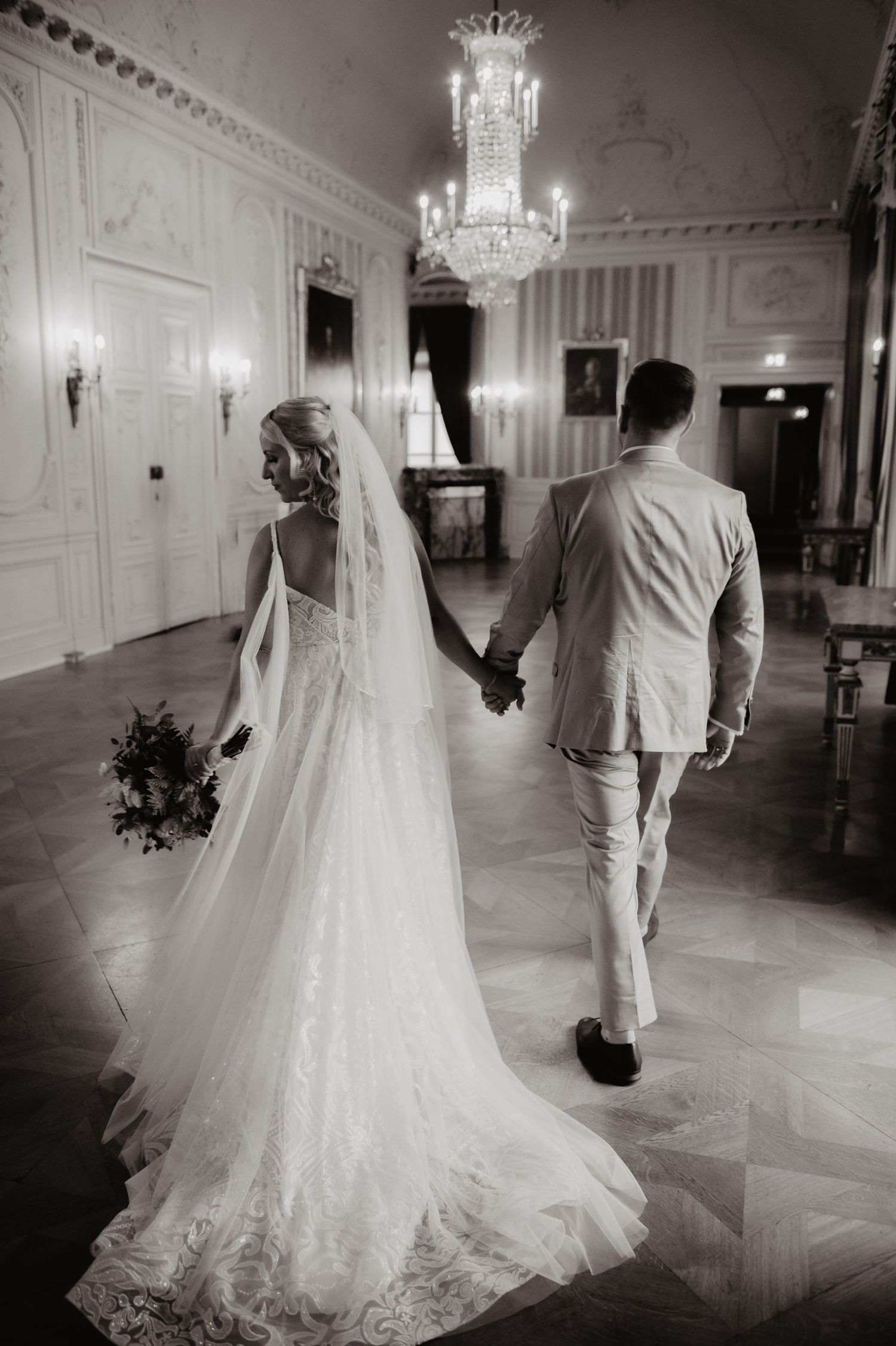 Black and white photo of bride and groom holding hands walking in elegant ballroom with crystal chandelier.