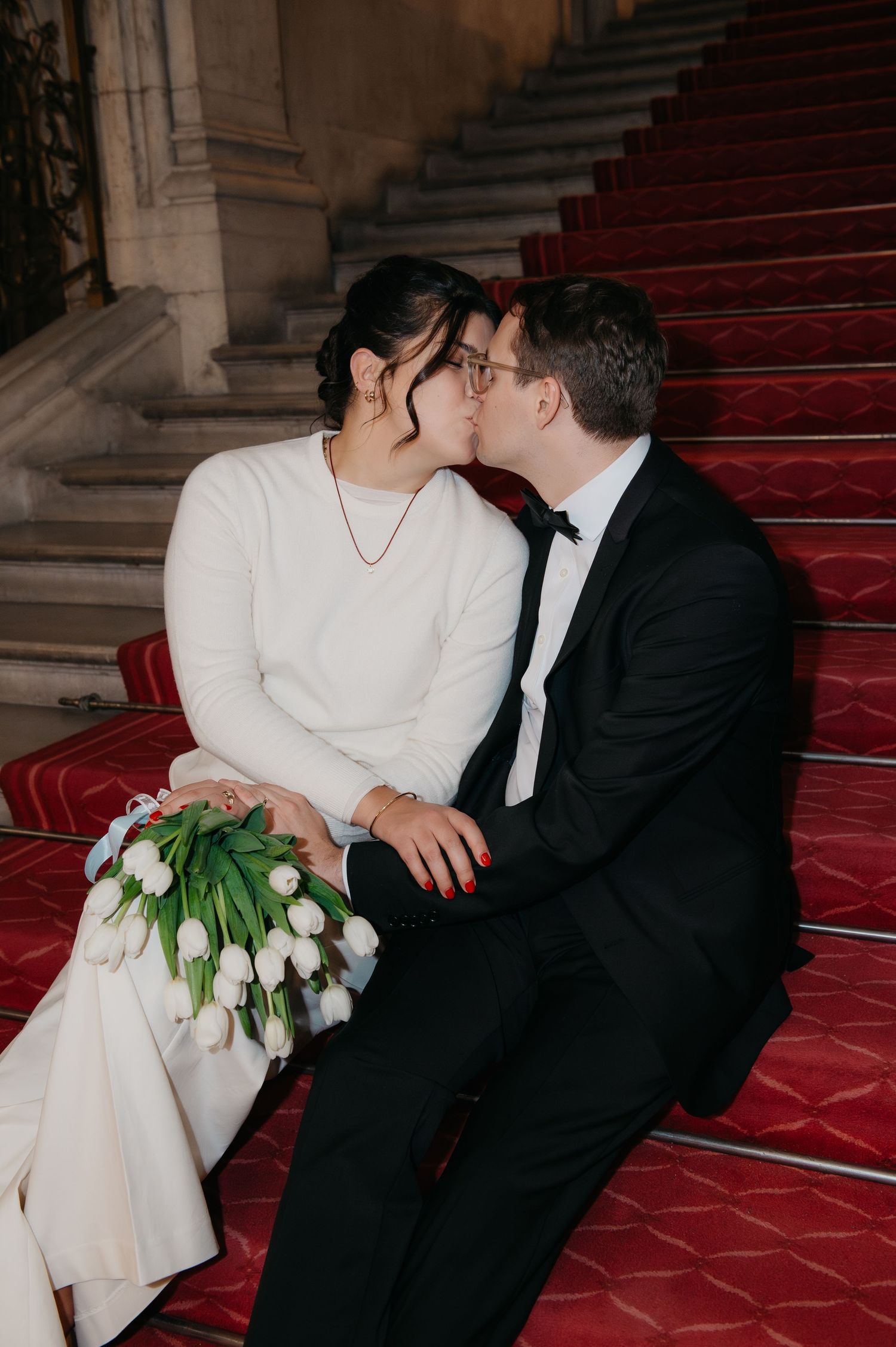 Newlywed couple sharing a kiss on red carpeted stairs, bride holding white tulips bouquet in elegant wedding attire.
