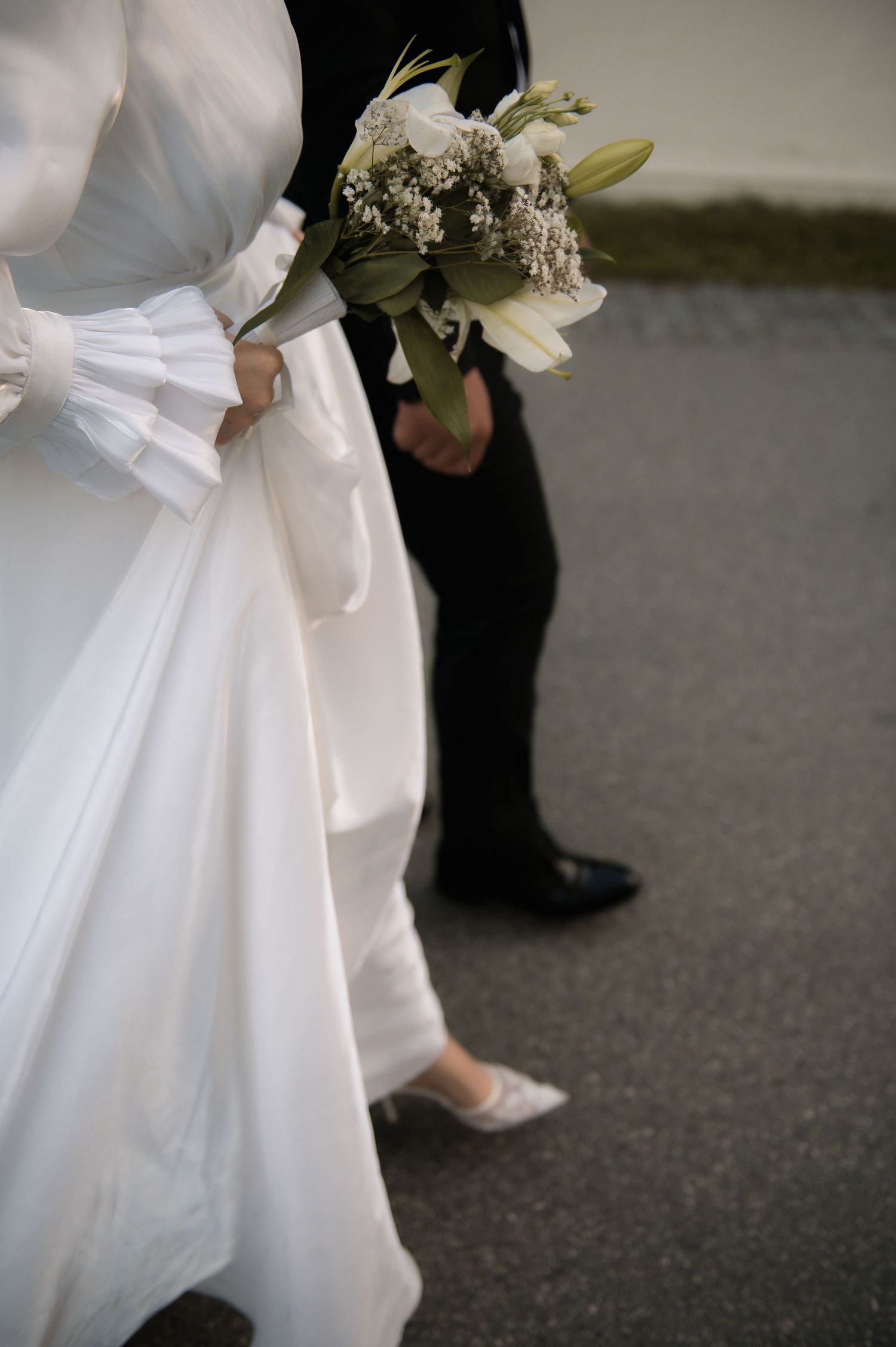 Bride in white gown holding bouquet of white lilies stands beside groom in black suit on pavement.