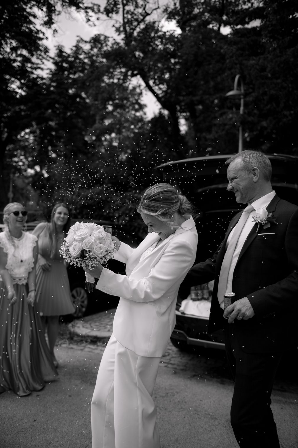 Black and white photo of bride in white suit holding bouquet while being showered with confetti outside.