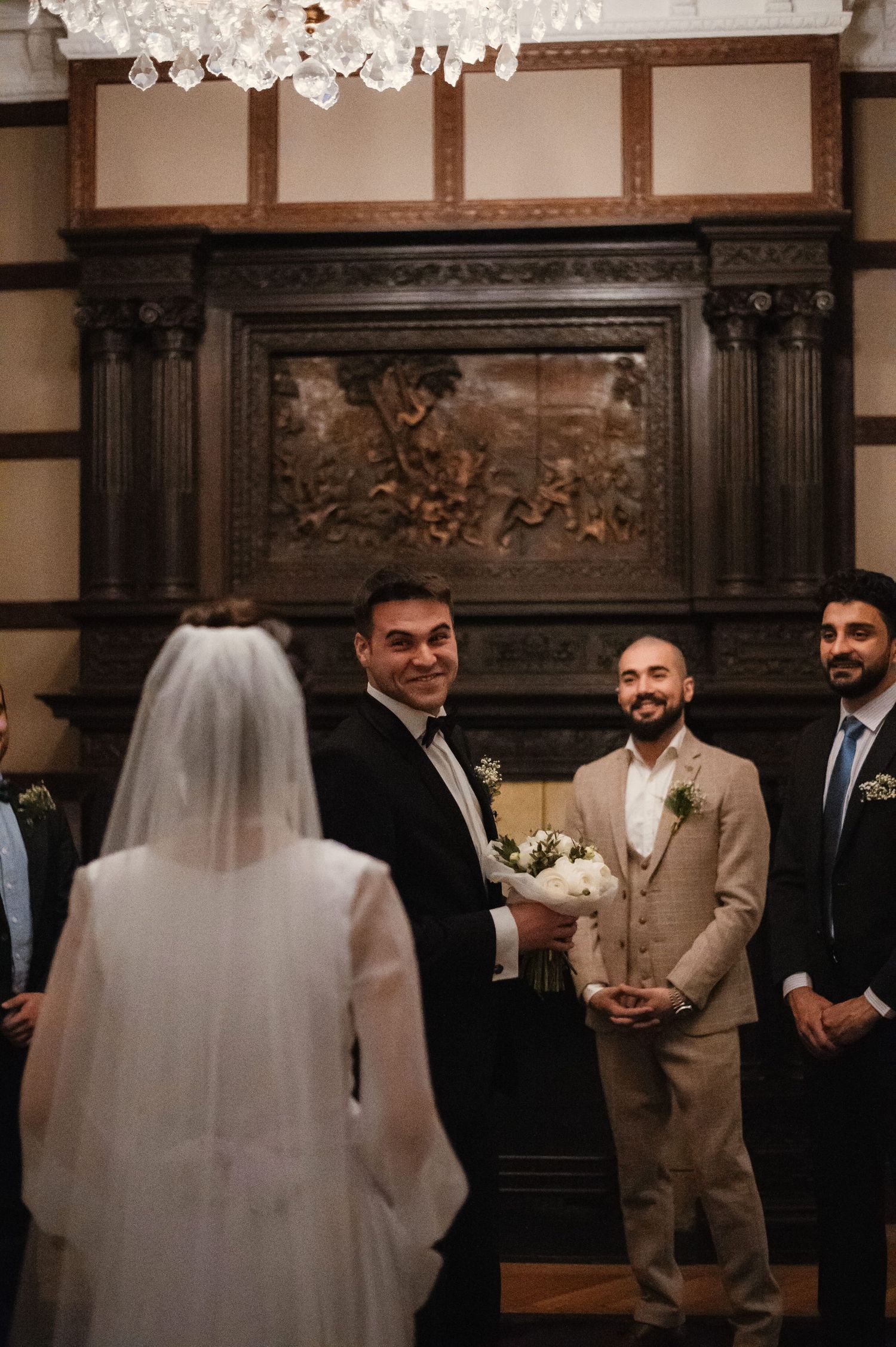 Groom smiling at bride in white veil during indoor wedding ceremony beside ornate carved fireplace.