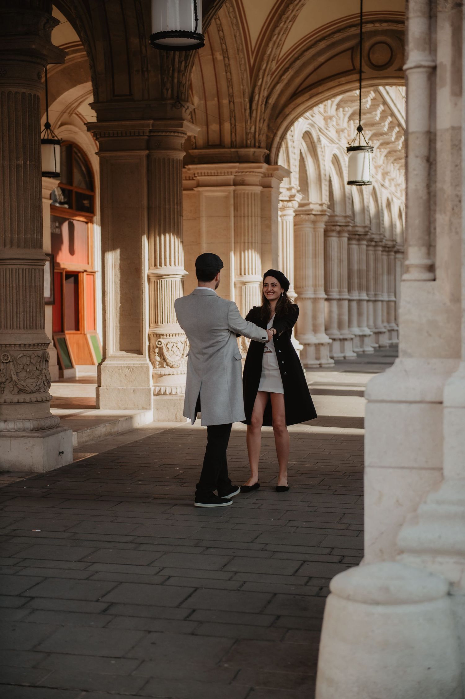 Elegant couple holding hands and smiling in a sunlit classical European arcade with ornate columns.