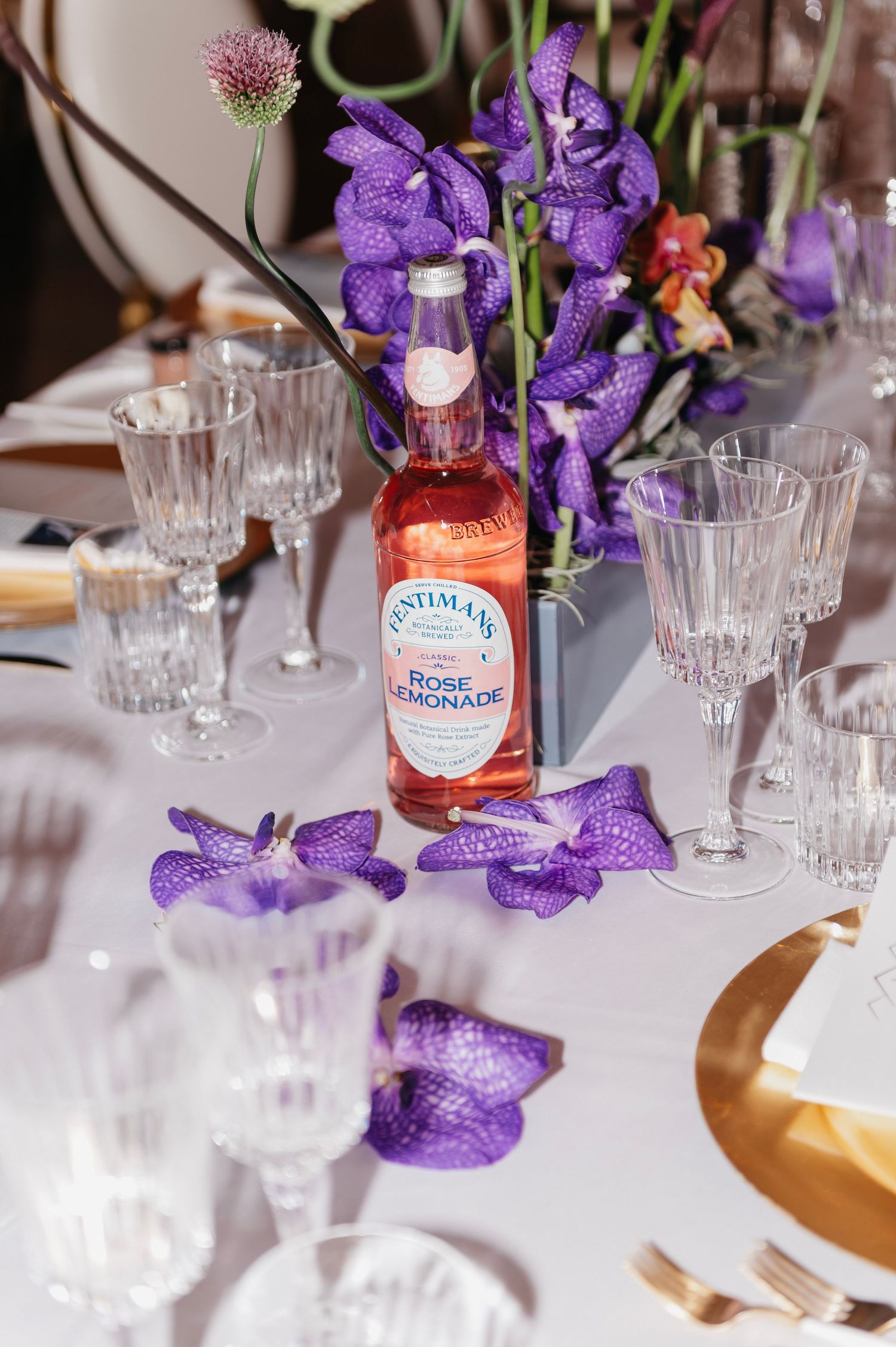 Elegant table setting with Fentimans Rose Lemonade, crystal glasses, gold plates, and purple orchid floral centerpiece.