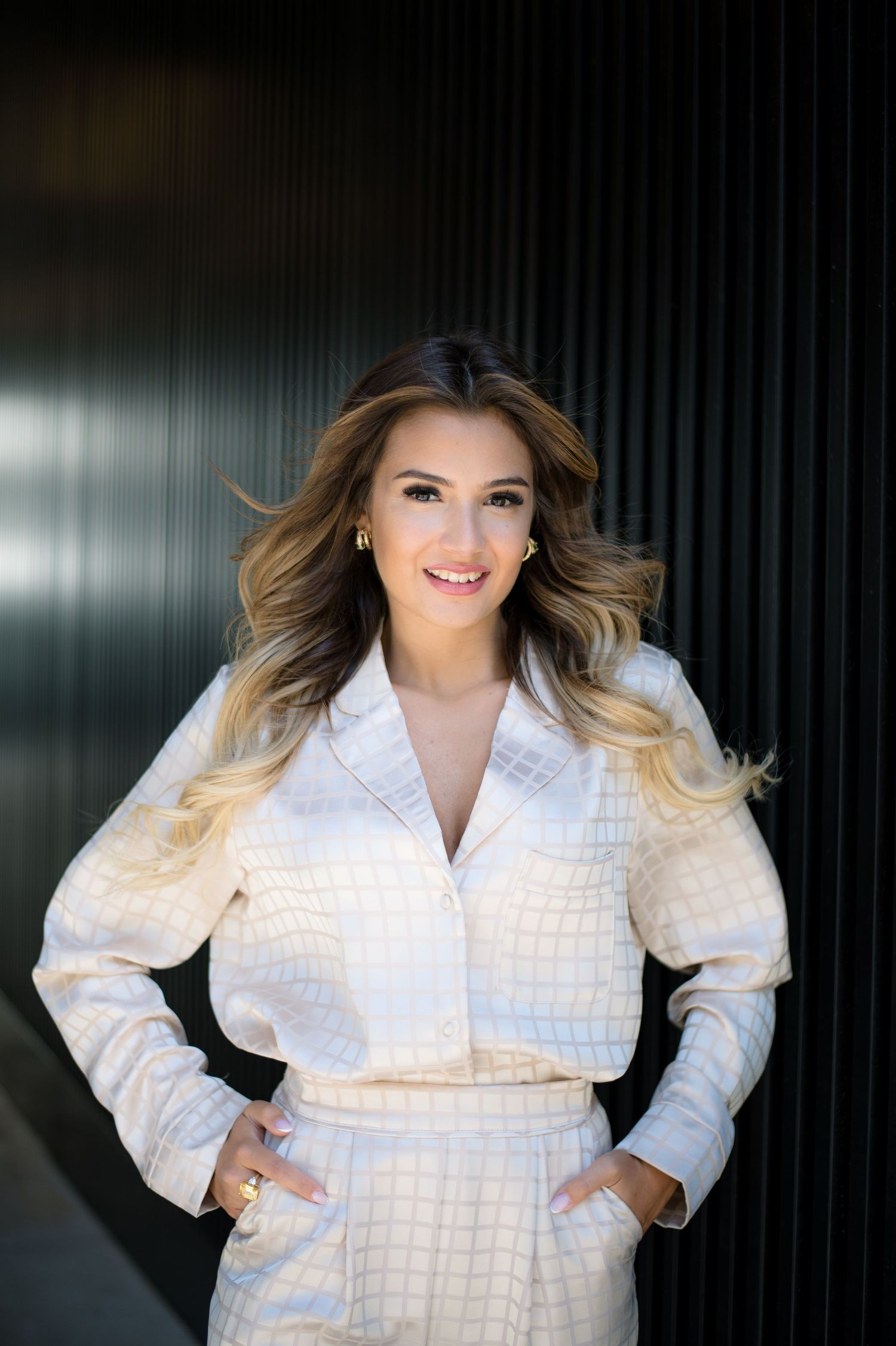 Woman in white textured co-ord suit smiling confidently with hands on hips against dark background.